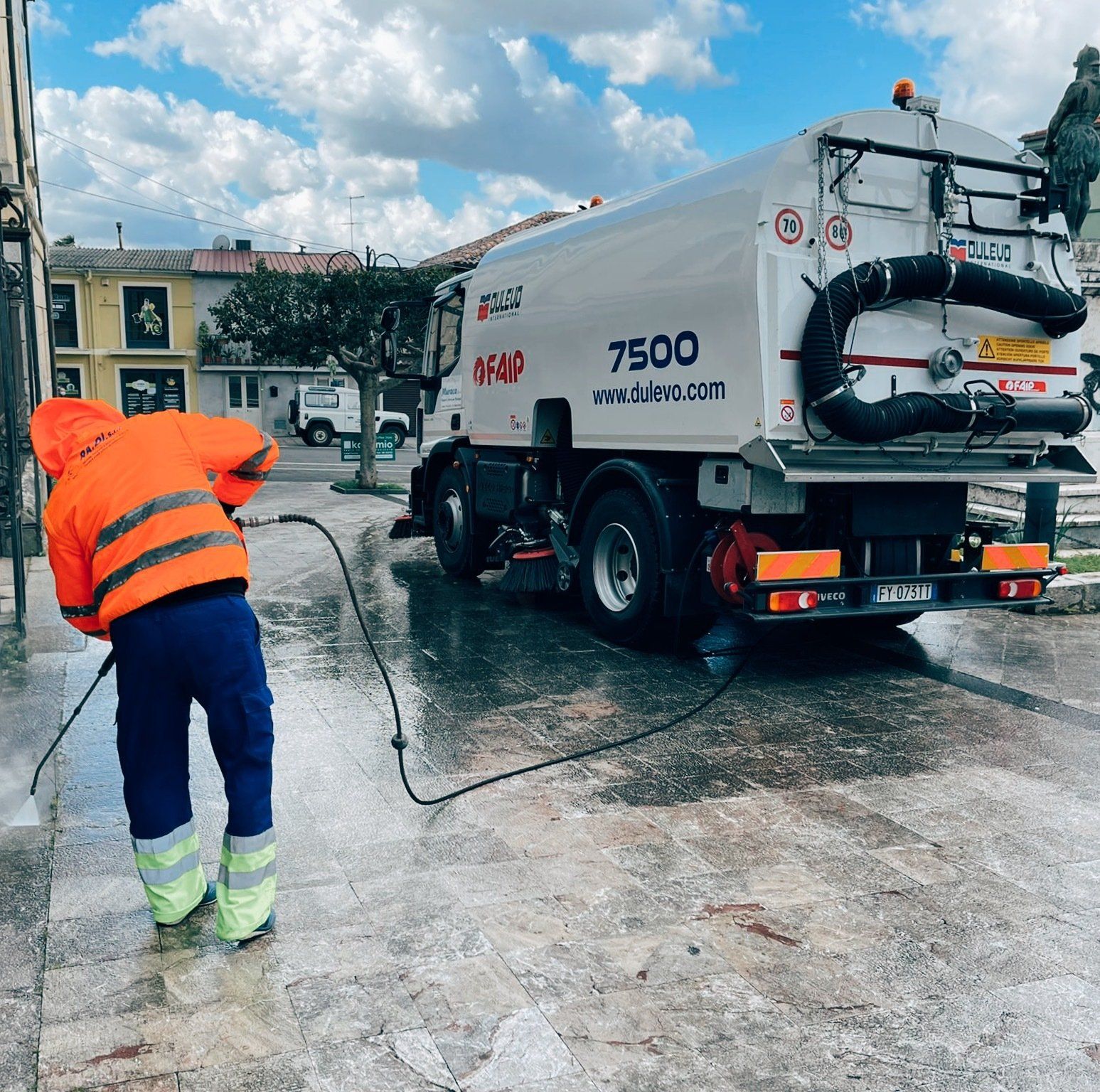 Un operaio con la giacca arancione pulisce una strada con un camion aspiratore bianco sotto un cielo nuvoloso.