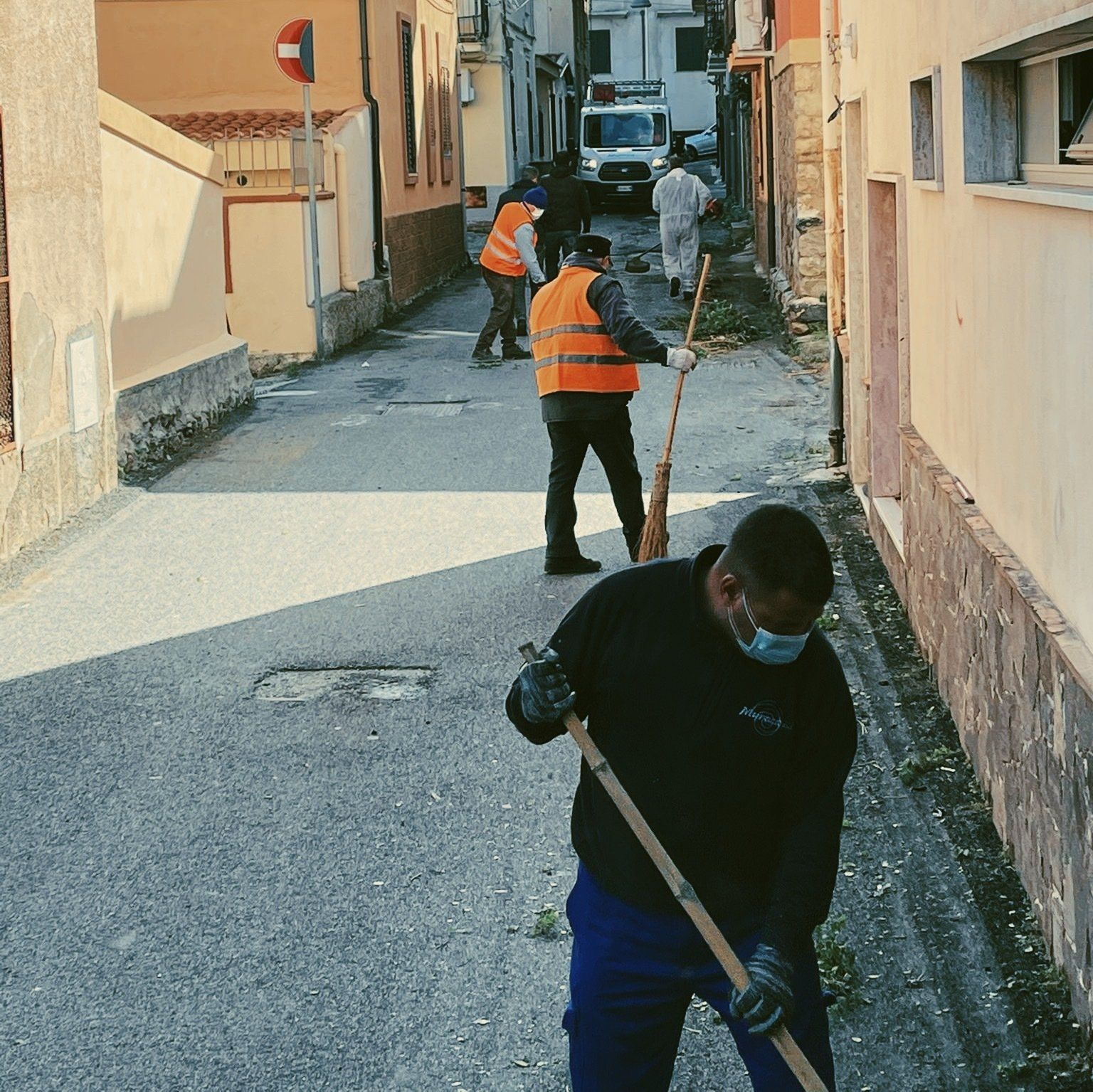 Operai intenti a pulire una strada stretta, uno dei quali spala in primo piano, mentre altri rimuovono i detriti più avanti.