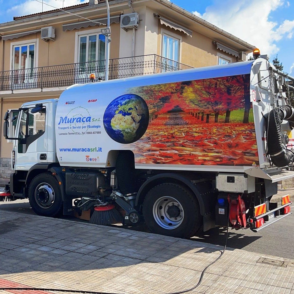 Un camion della spazzatura bianco con un colorato murale della Terra è parcheggiato in una strada accanto a un edificio beige.
