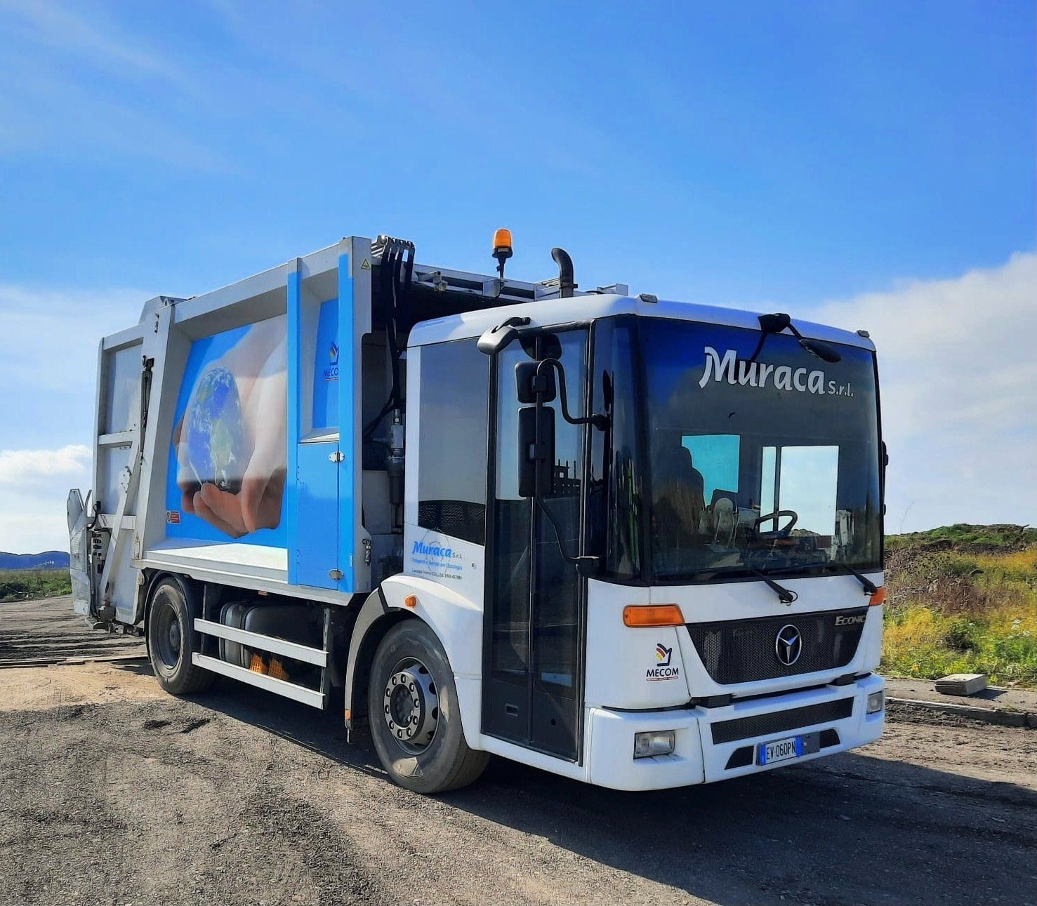 Un camion della spazzatura bianco con pannelli laterali blu parcheggiato su una strada sterrata sotto un cielo azzurro e limpido.