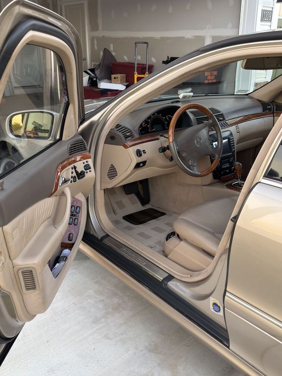 Beige vintage car interior with open driver’s door in a garage, showing cream seats and wood steering wheel