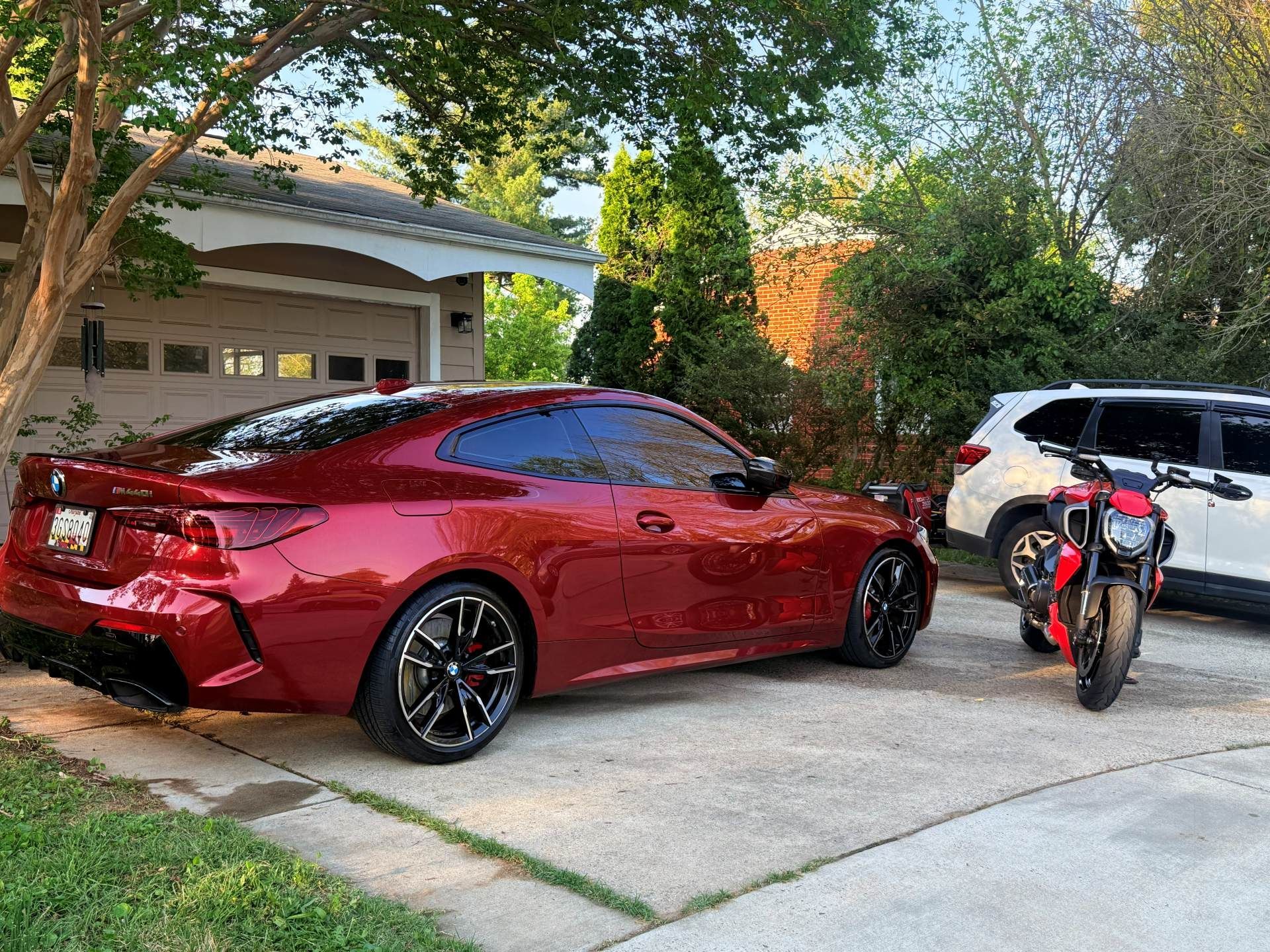 Red sports coupe and a red dirt bike parked in a driveway beside a garage and trees.