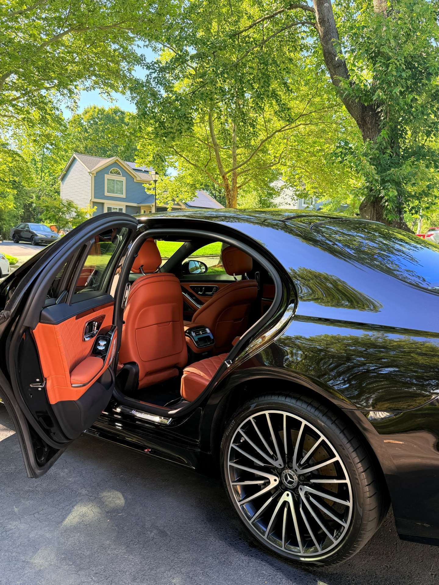 Black luxury sedan with orange leather interior, rear door open on a tree-lined suburban street.