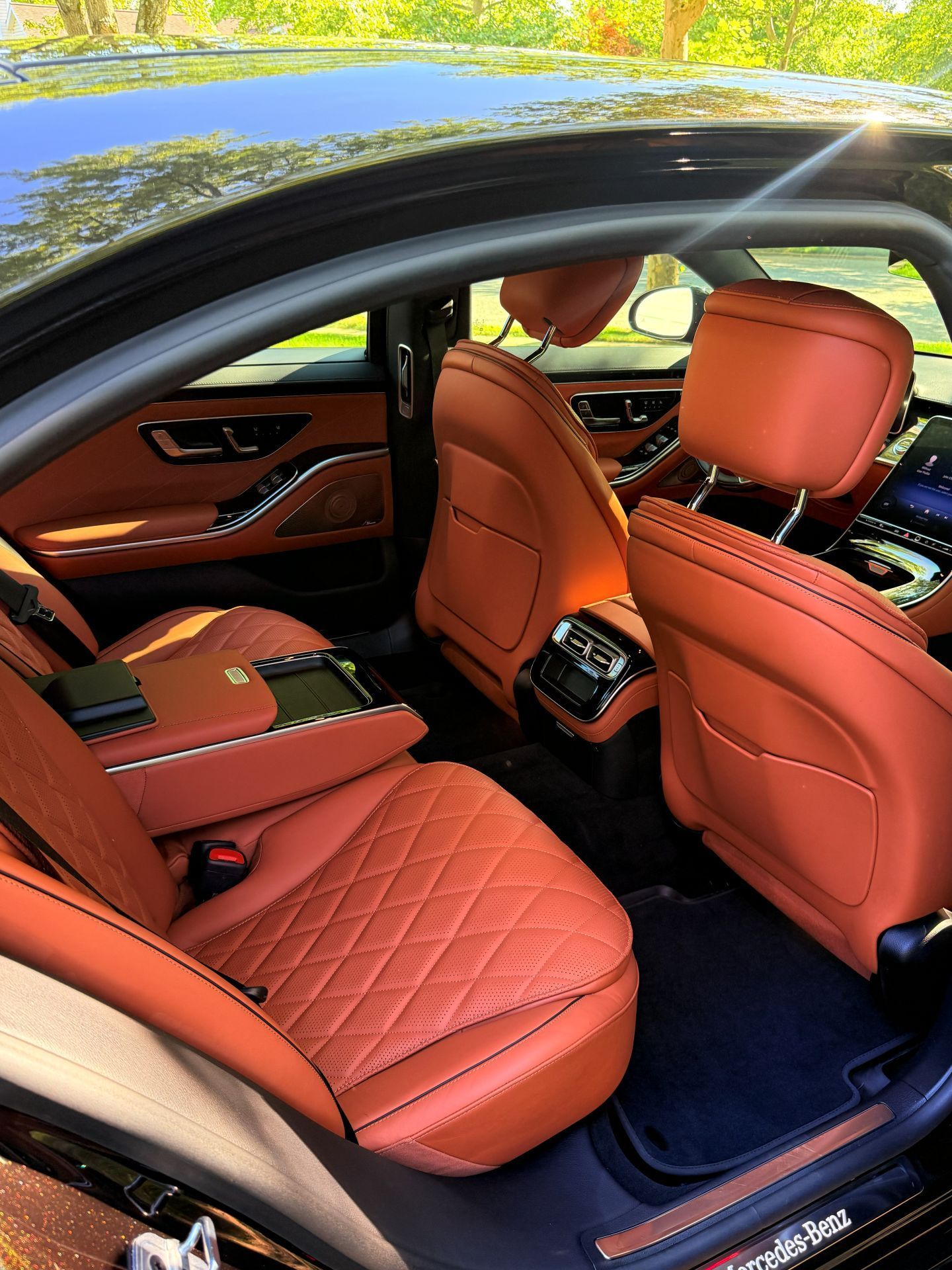 Luxury car interior with orange quilted leather seats and open door, parked by water and trees.