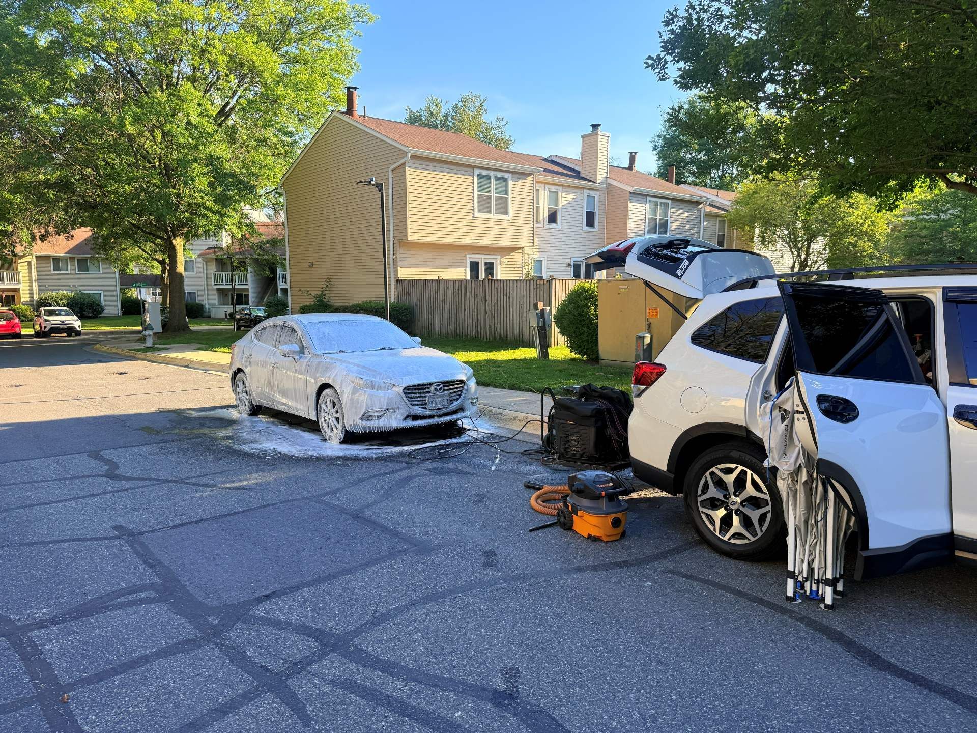Damaged white SUV with open hatch and tools beside it on a suburban street; white sedan parked nearby.