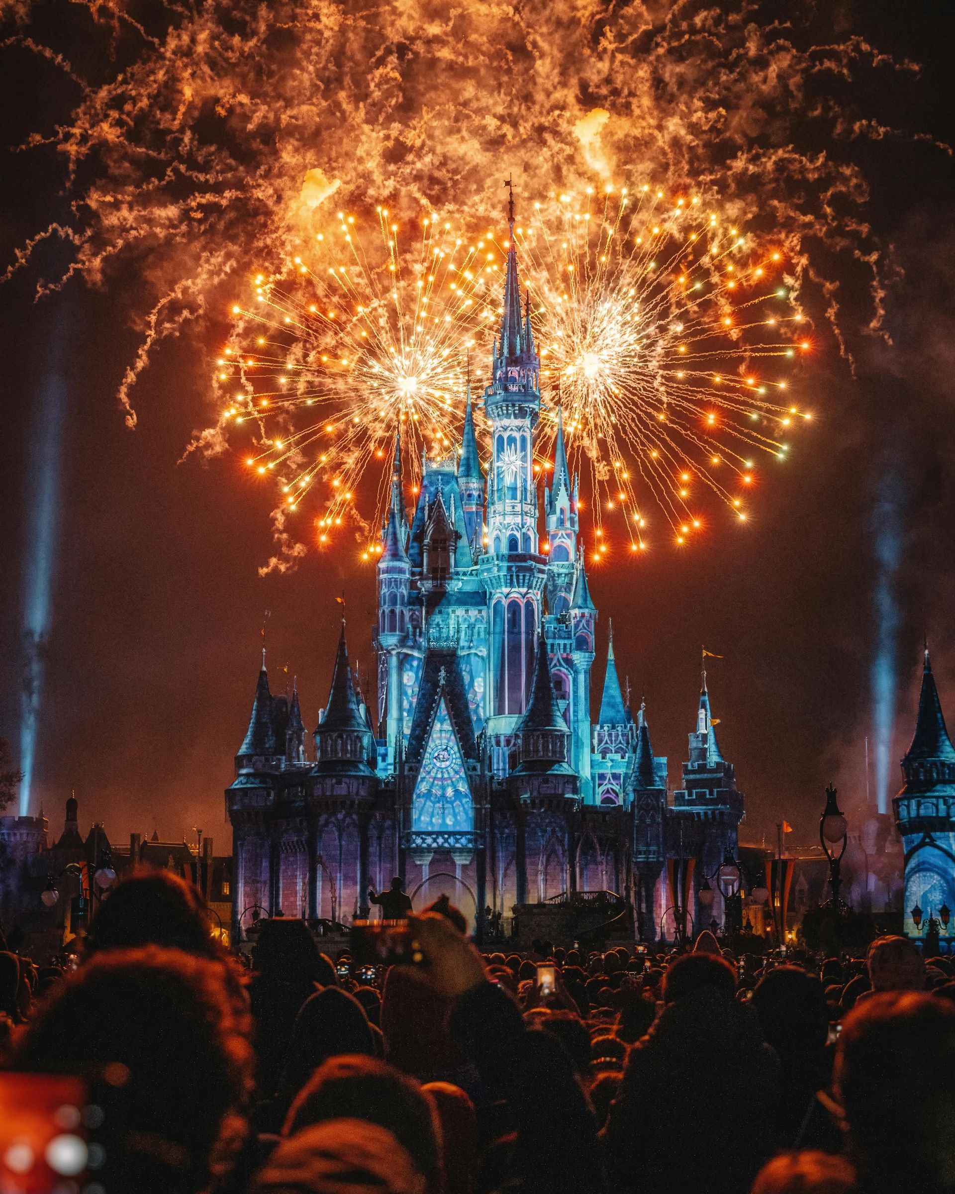 Fireworks explode in vibrant orange hues above a tall, illuminated Disney castle as a large crowd watches at night.