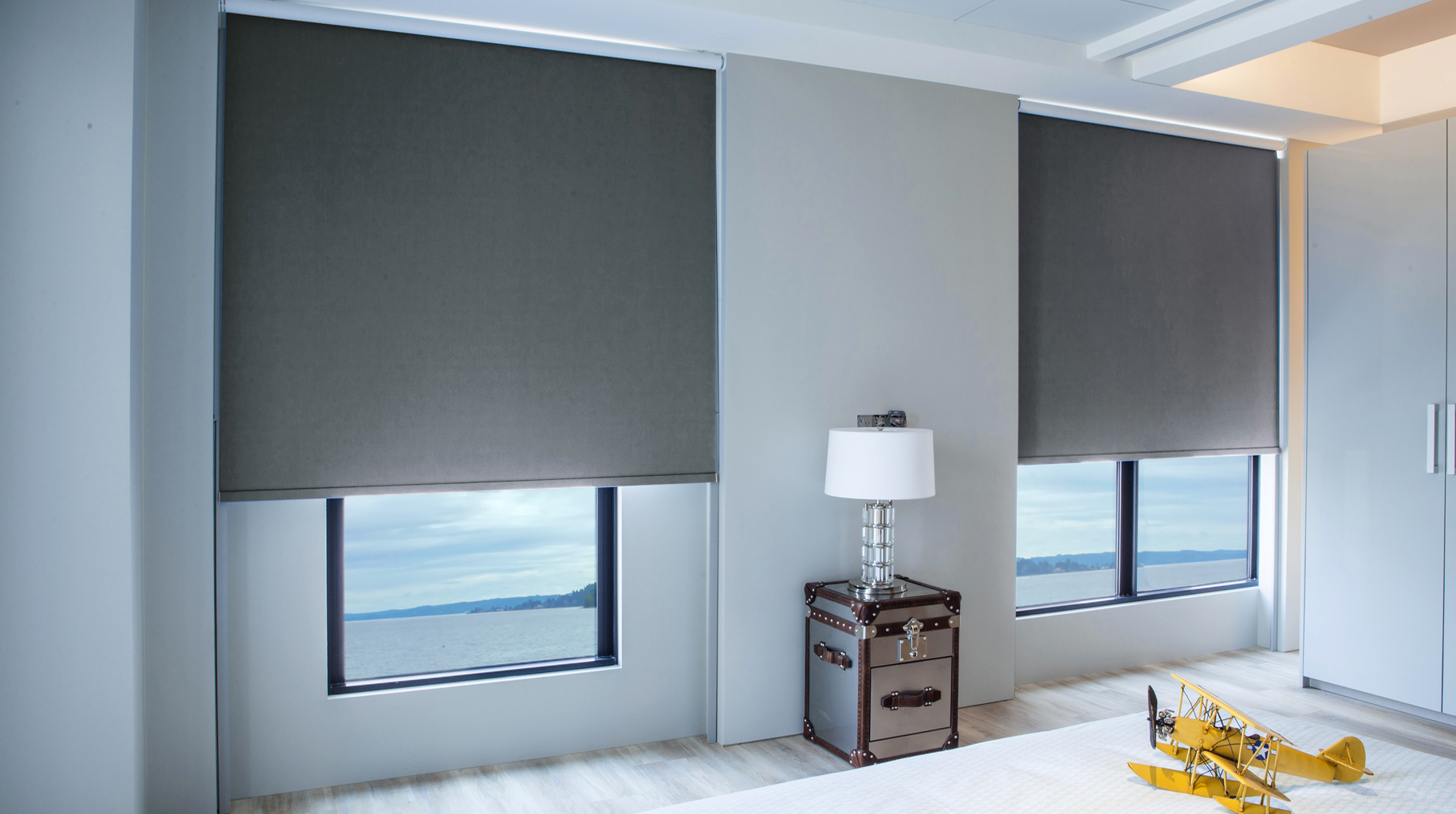 a bedroom with two windows , a lamp , a trunk and a plane on the floor with roller shades All Window Decor (817) 448-3393.