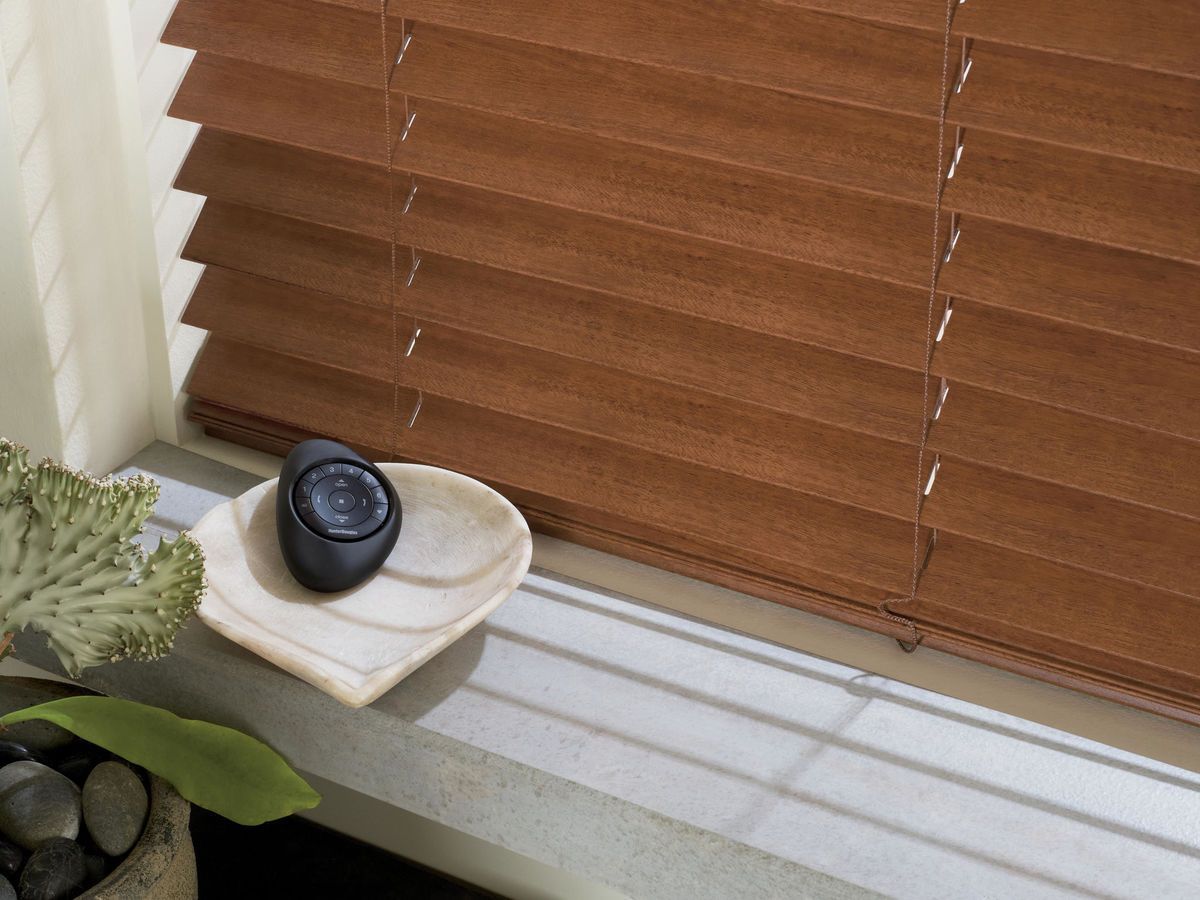a motorized wooden blind with a remote sitting on a window sill next to a plant All Window Decor (817) 448-3393