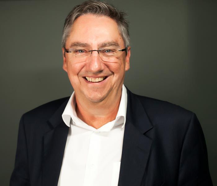 Smiling man in a dark blazer and light shirt against a blue gradient background