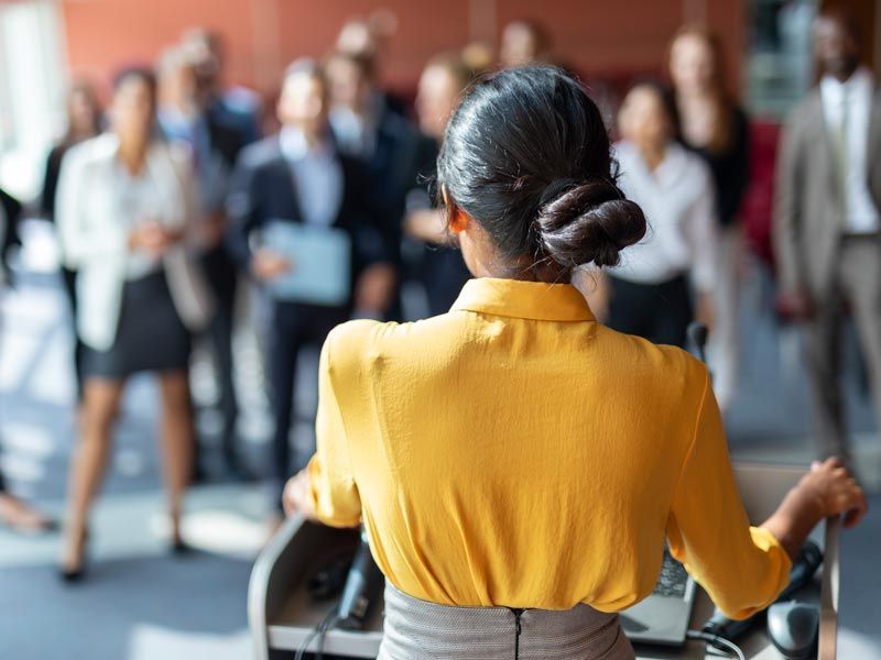 Person in a yellow shirt addressing a blurred audience in a conference room