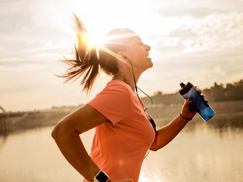 Woman jogging outdoors at sunrise, holding a water bottle and wearing a coral shirt