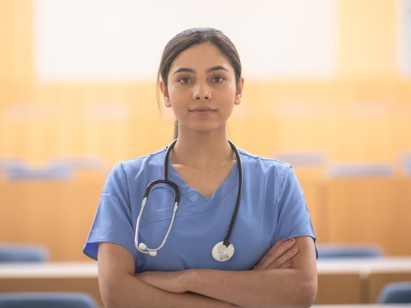 Nurse in blue scrubs with stethoscope stands with arms crossed in a bright classroom or lecture hall.