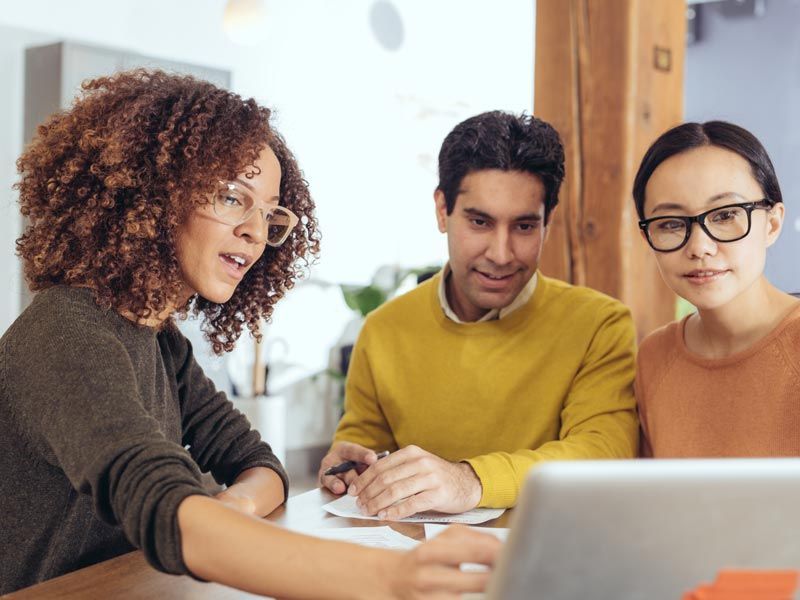 Three colleagues discussing papers at a table with a laptop in a bright office