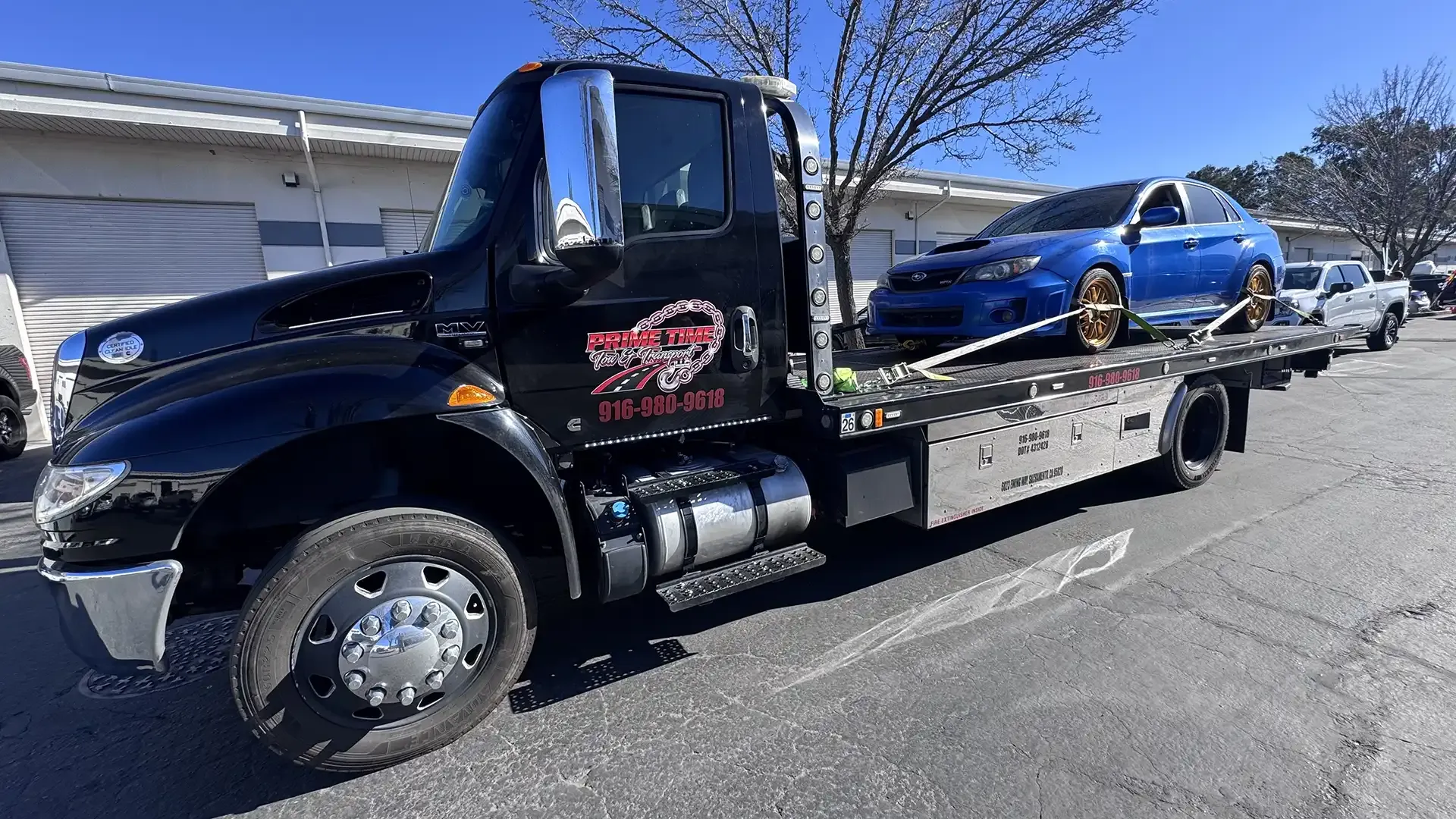 A black tow truck with a blue car on the back is parked on the side of the road.