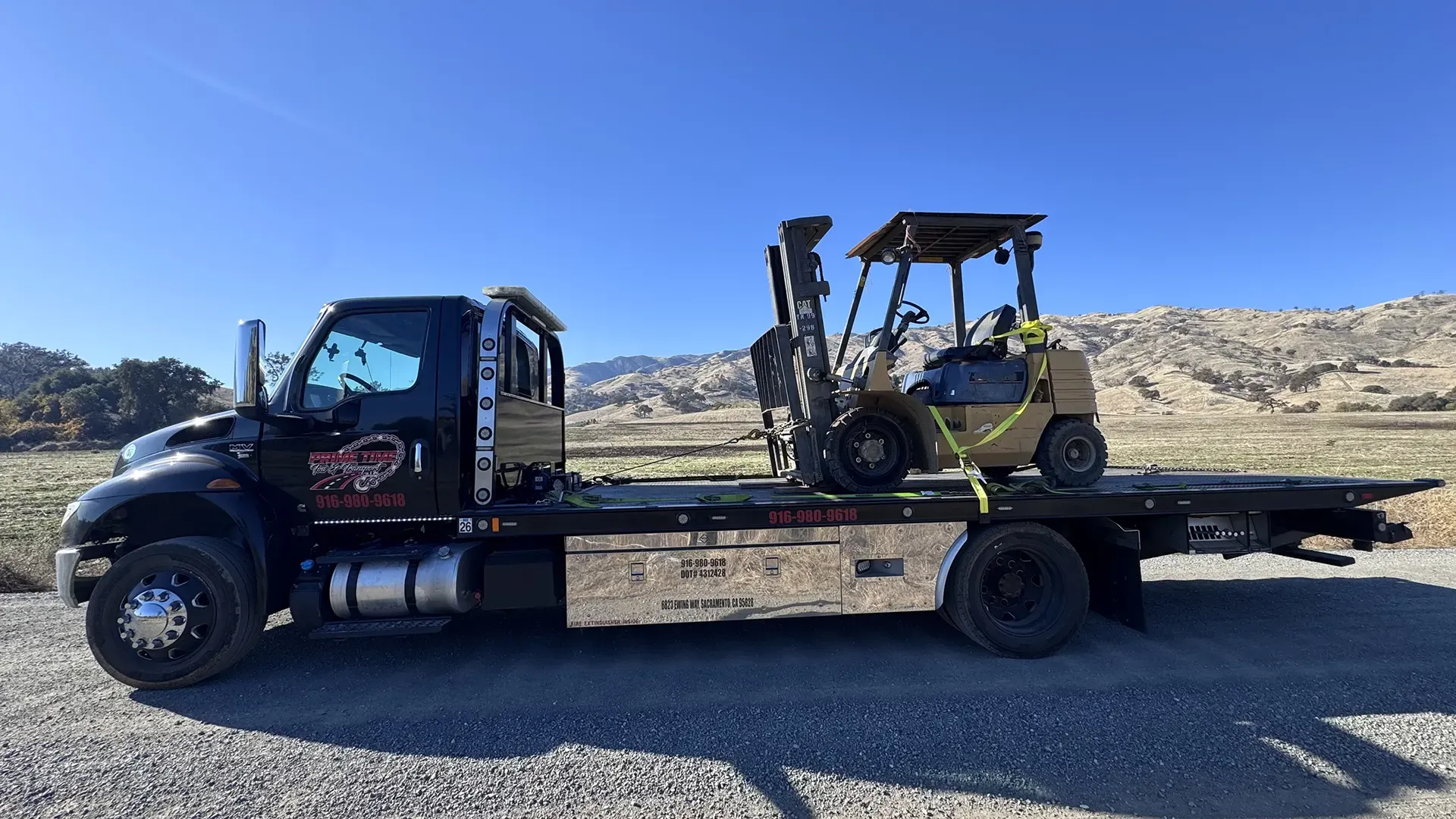 A tow truck is carrying a forklift on the back of it.