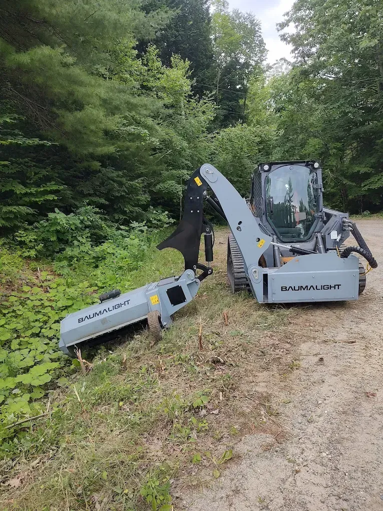 Compact track loader with a mower attachment working beside a wooded roadside.