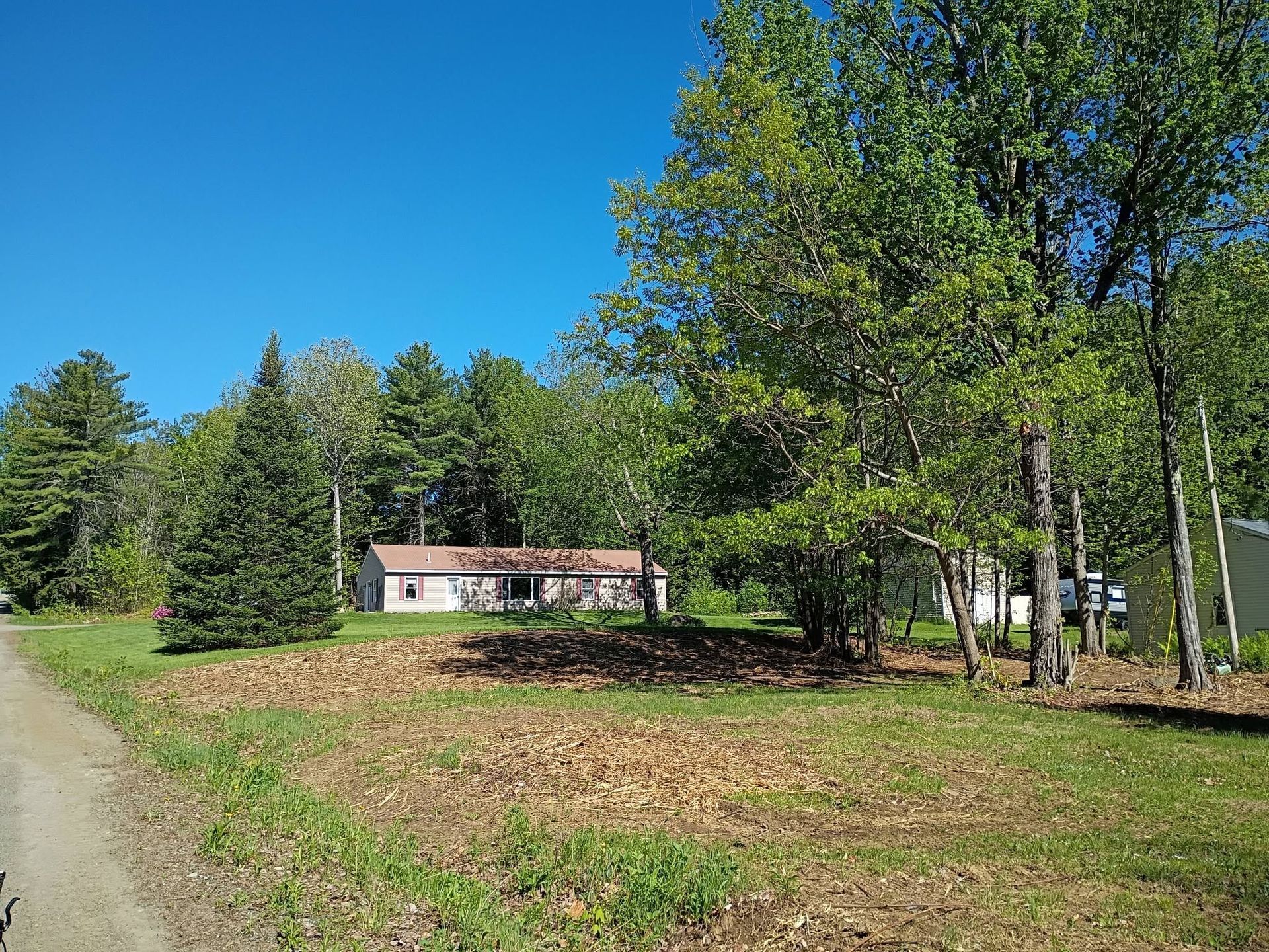 Rural house by a dirt road, surrounded by green trees and open grassy field under a blue sky