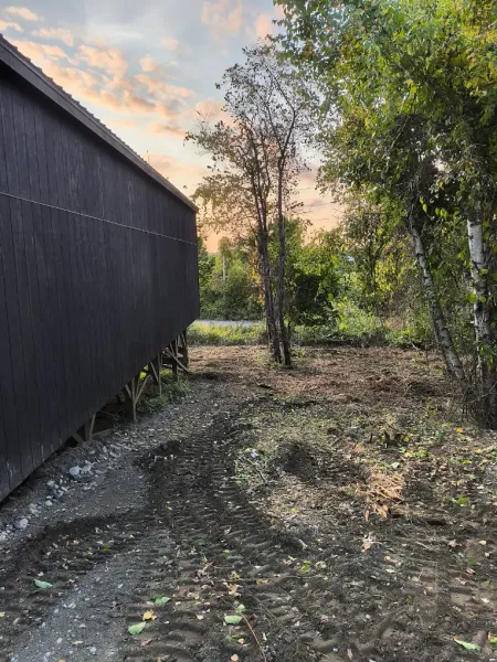 Narrow dirt path beside a dark building, with trees and a grassy yard at sunset.