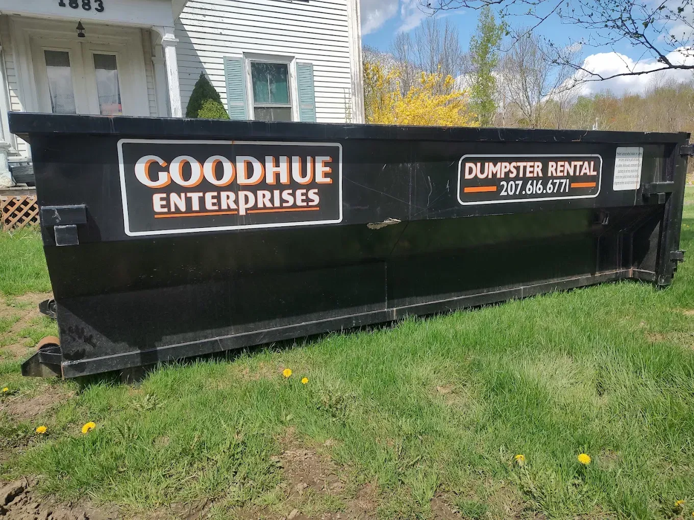 Black dumpster in grass beside house, labeled 