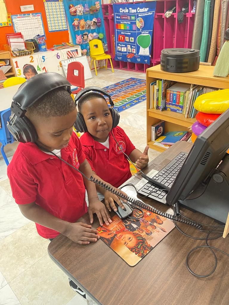 Two young boys wearing headphones are sitting at a desk in front of a computer.