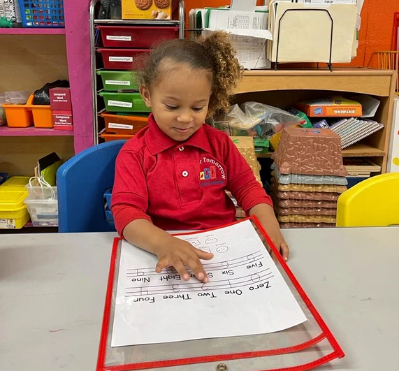 A little girl in a red shirt is sitting at a table looking at a piece of paper
