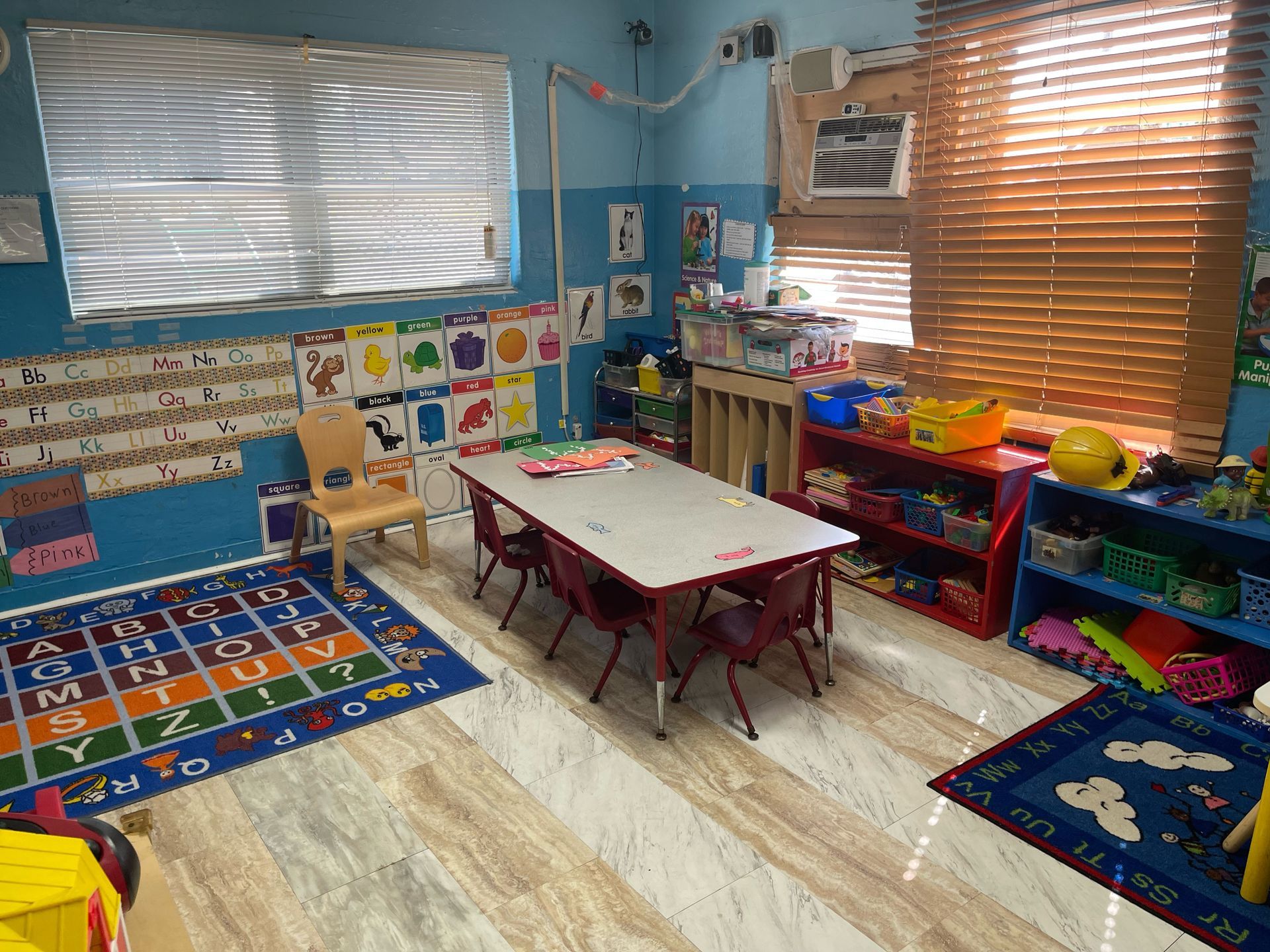 A classroom with a table and chairs and a rug with the alphabet on it.