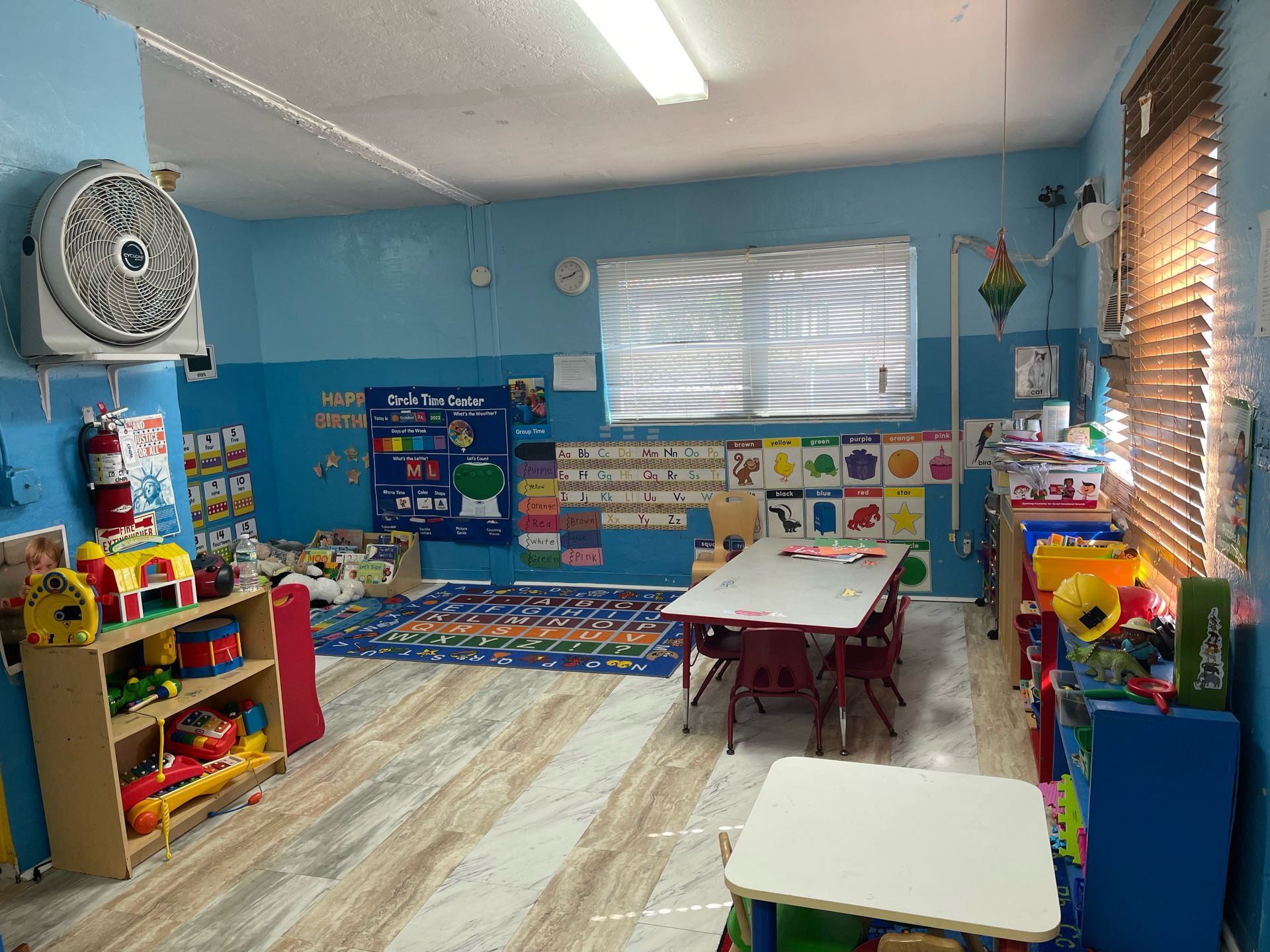 A classroom with tables and chairs and a fan on the wall.