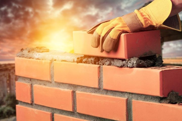 Gloved hand placing a brick on a sunlit brick wall with mortar against a bright sky