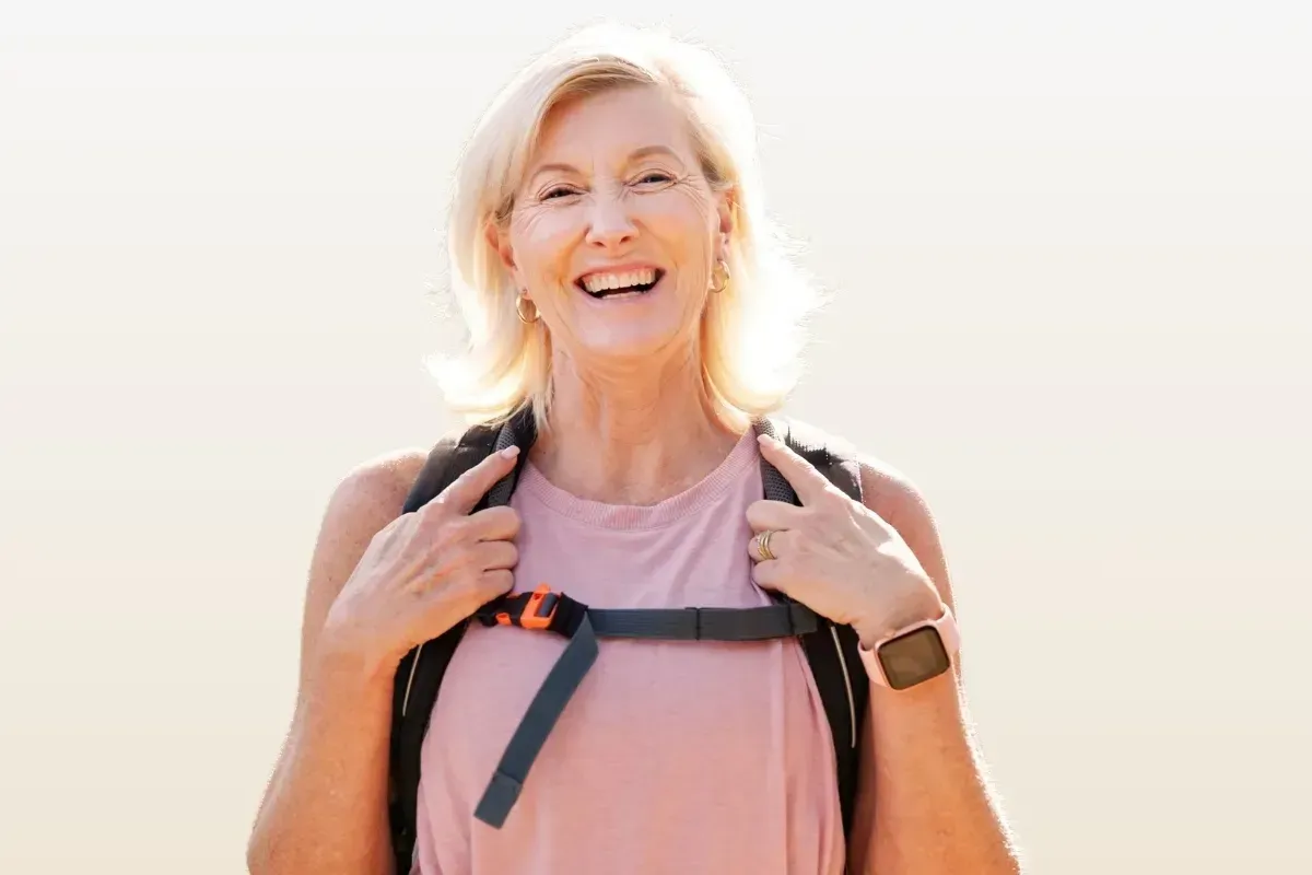 A woman wearing a pink tank top and a backpack is smiling.