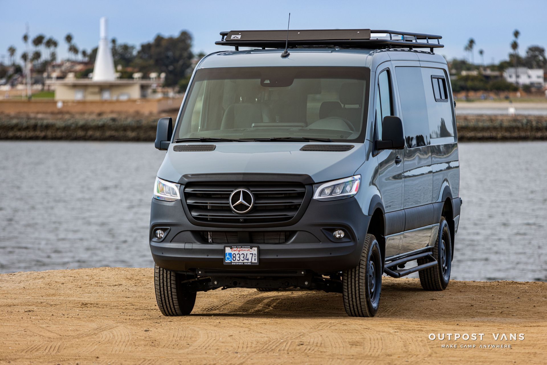 A mercedes sprinter van is parked on a sandy beach next to a body of water.
