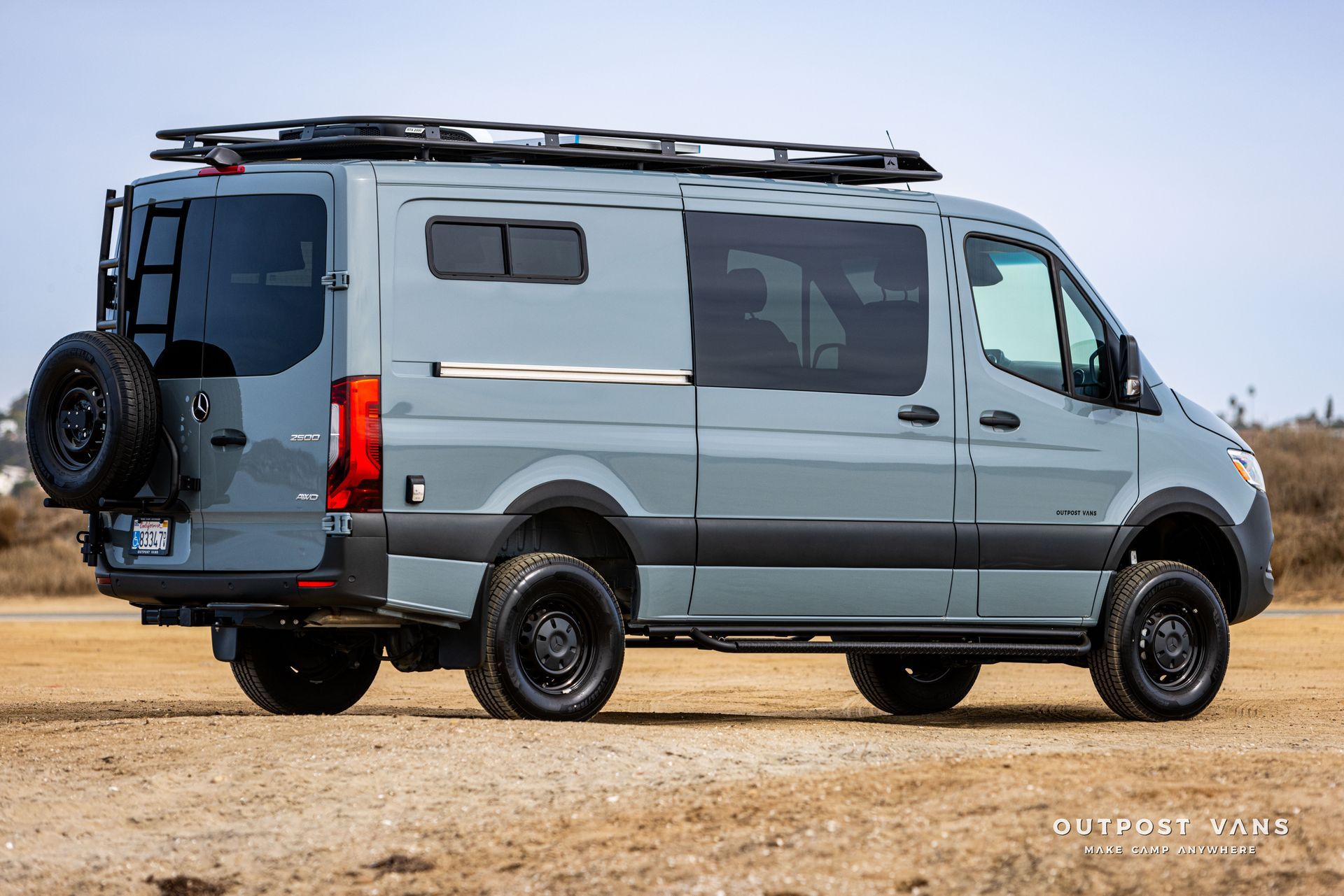 A gray van with a ladder on top of it is parked in a dirt field.