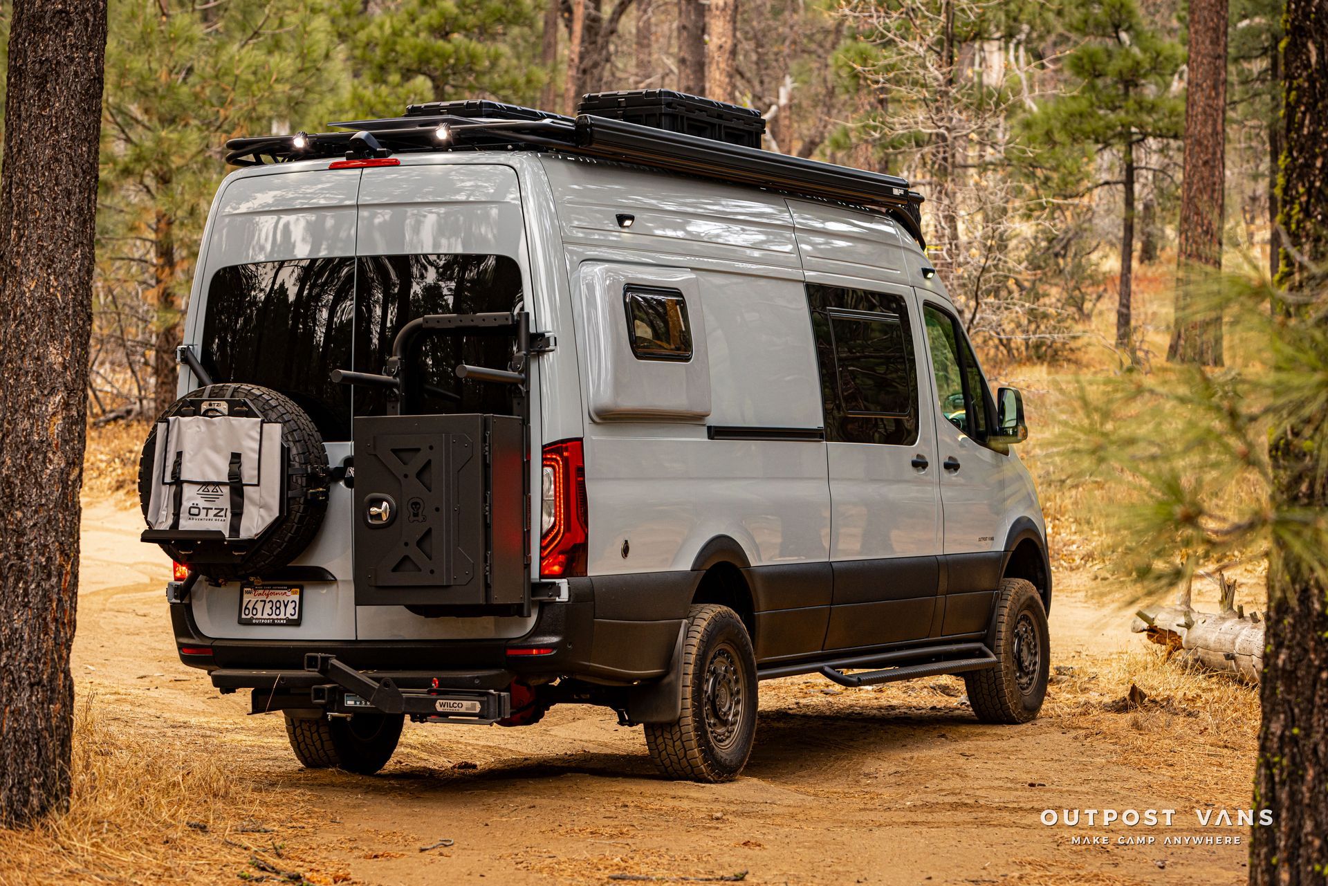 A gray van is parked on a dirt road in the woods.
