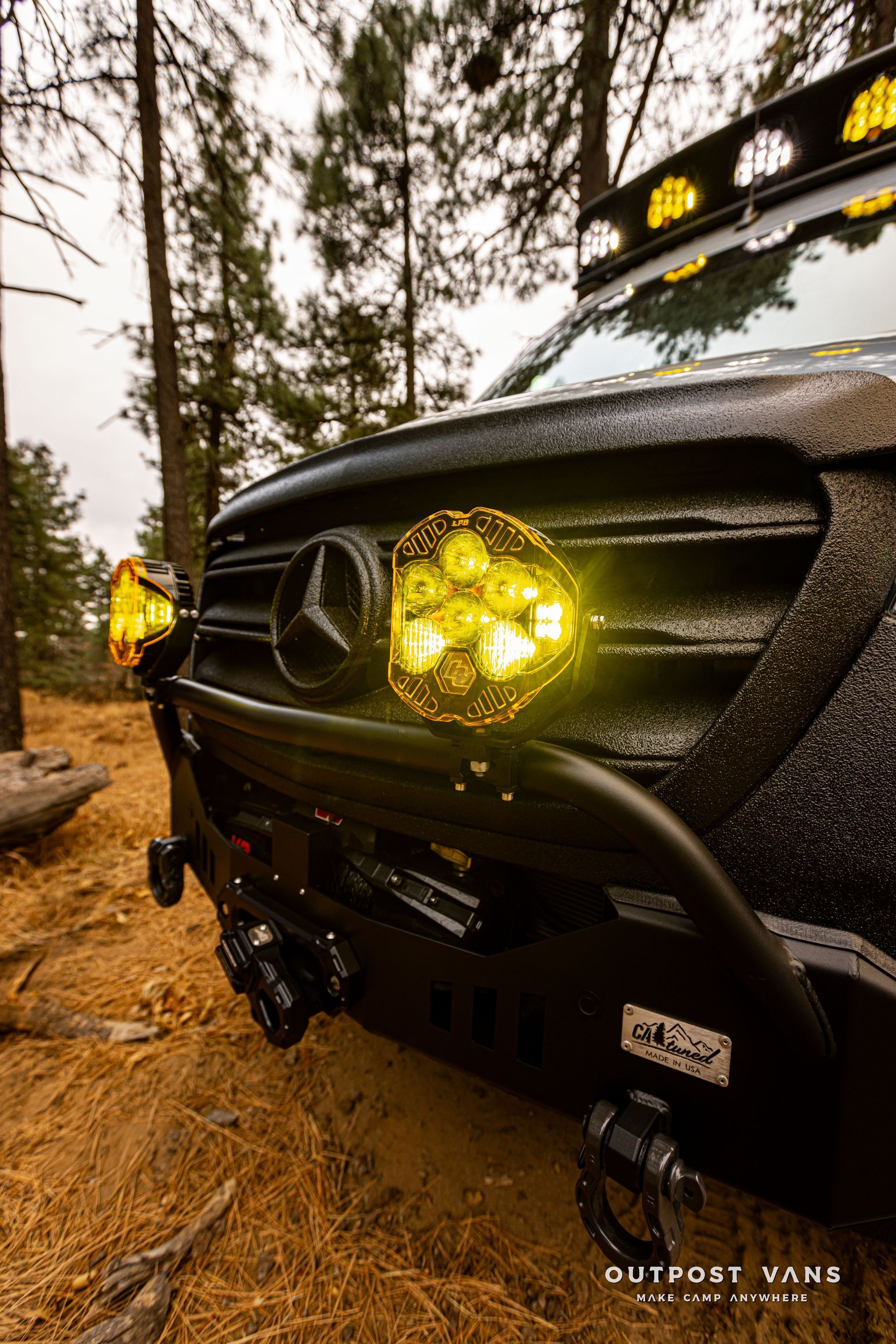 A black van with yellow lights on the front is parked in the woods.