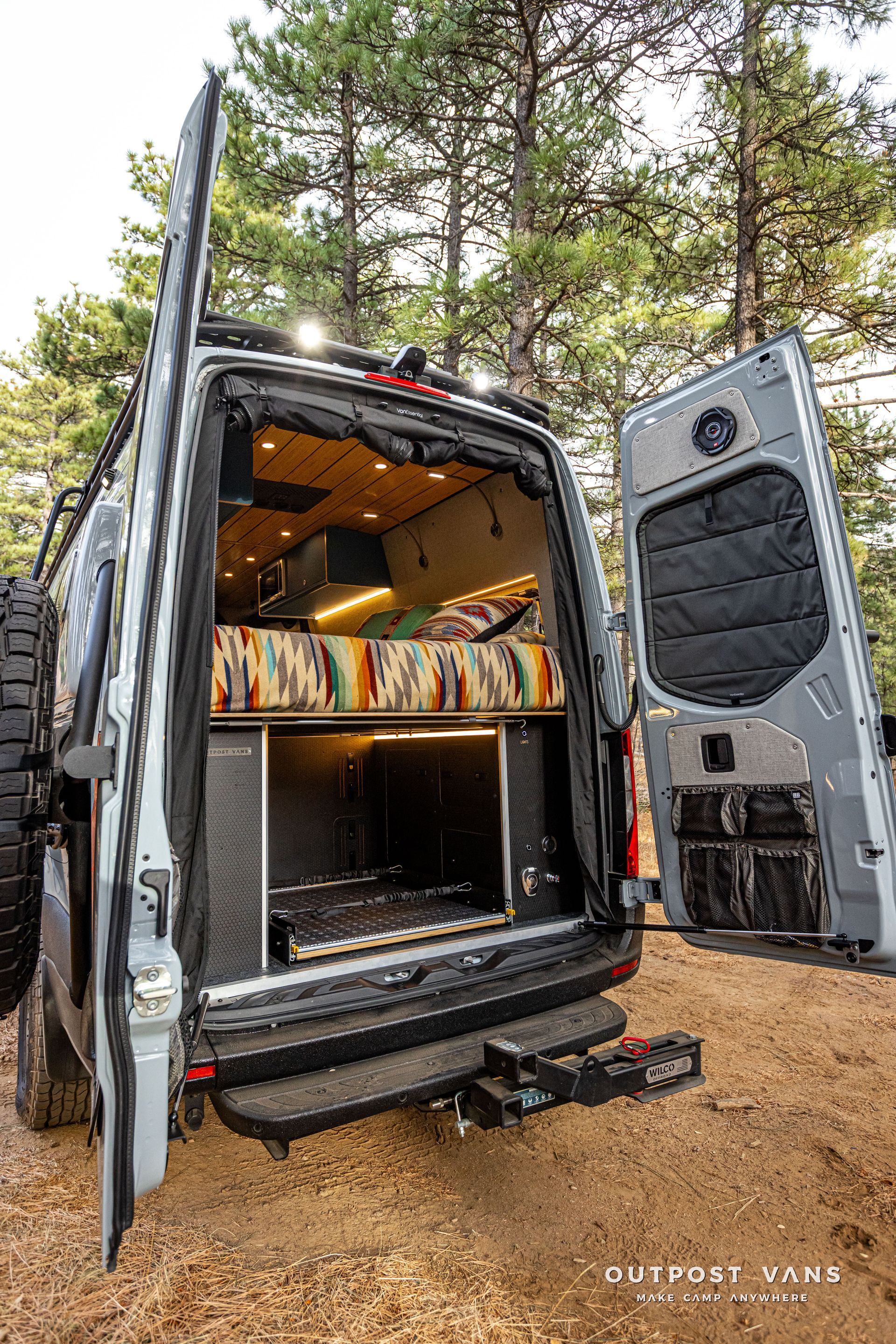 The back of a van with its doors open is parked in the woods.