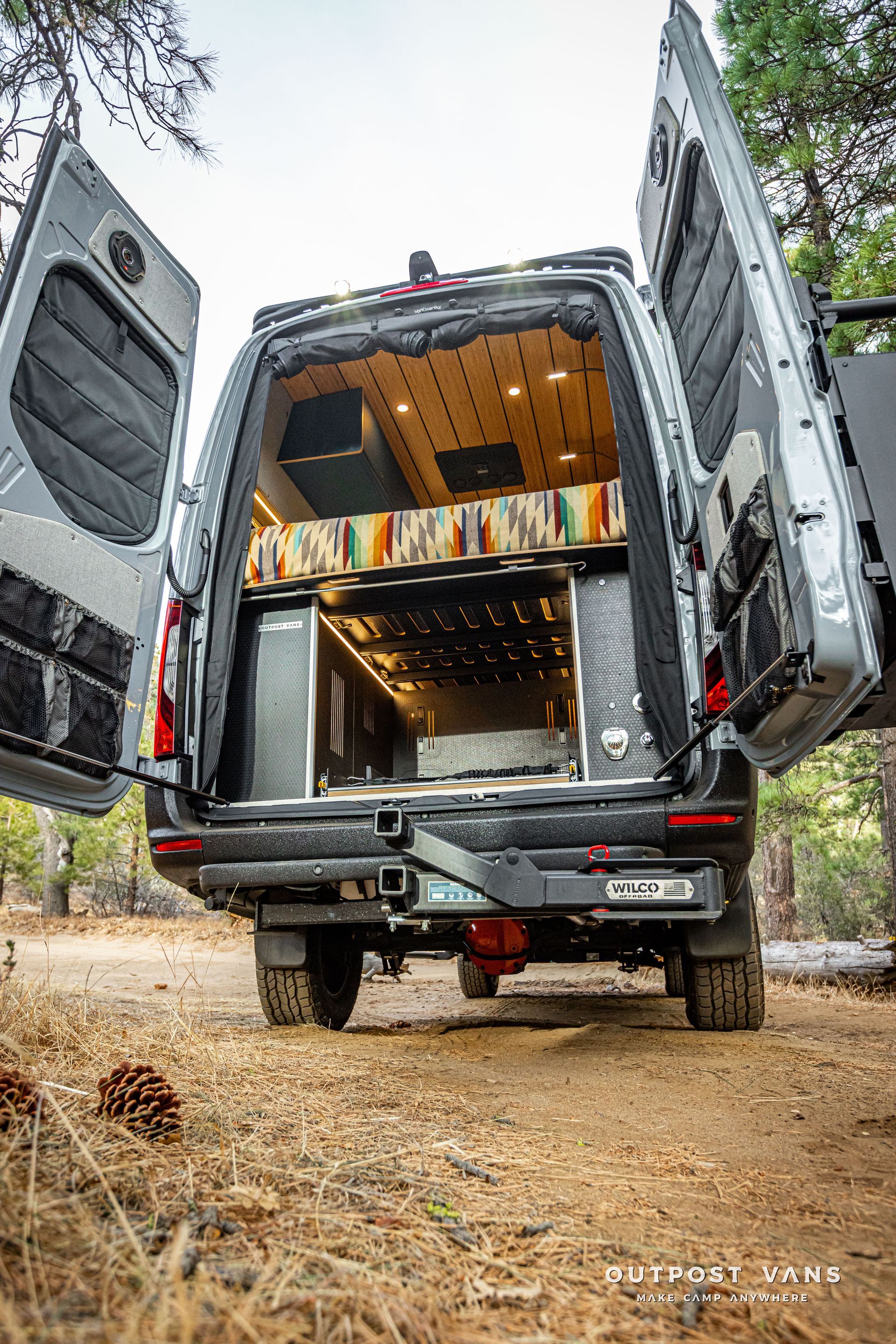 The back of a van with its doors open is parked in the dirt.