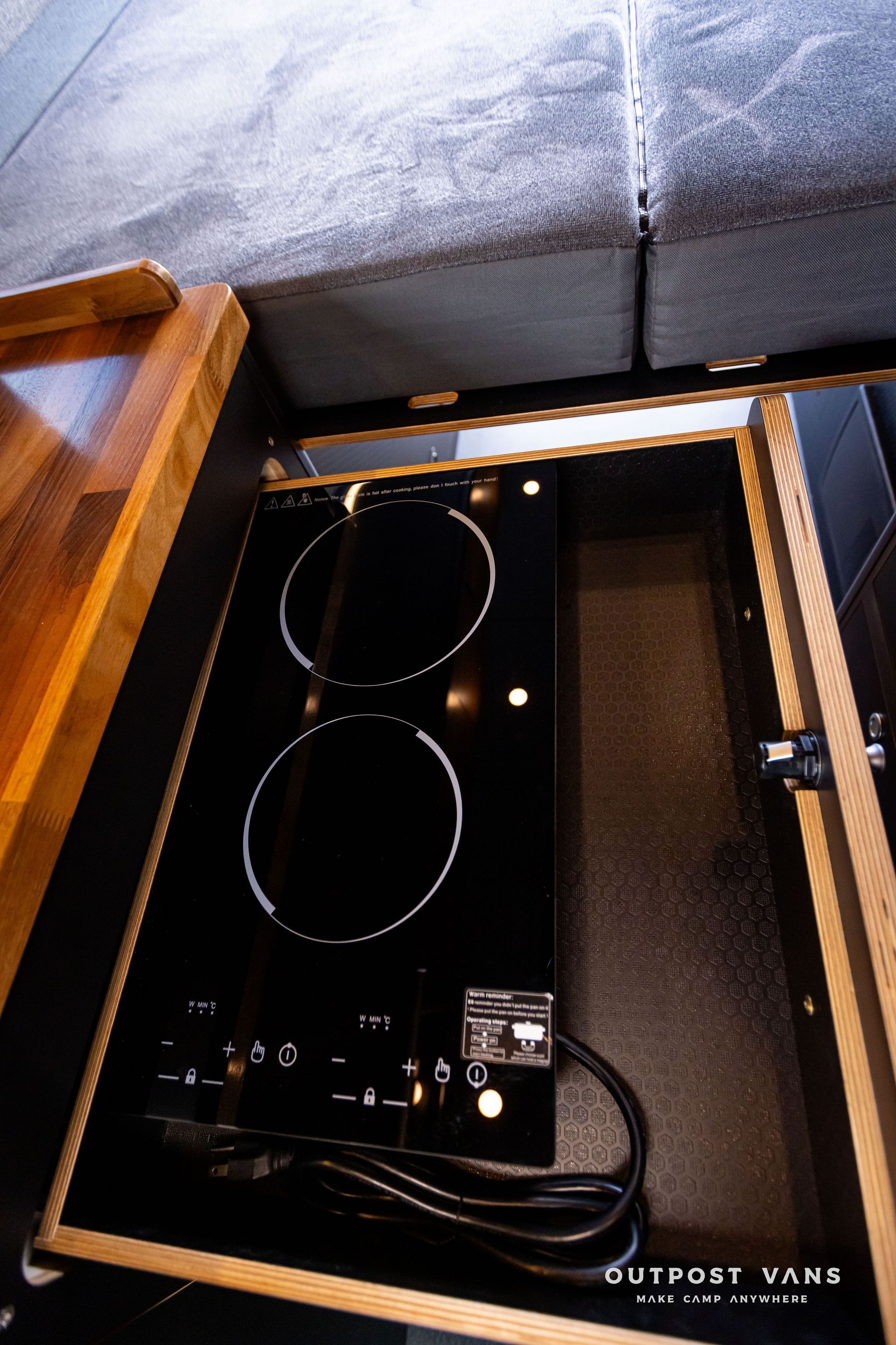 Outpost Vans custom built camper van Ford Transit 148 AWD. A two burner induction stove is sitting in a drawer under a bed.