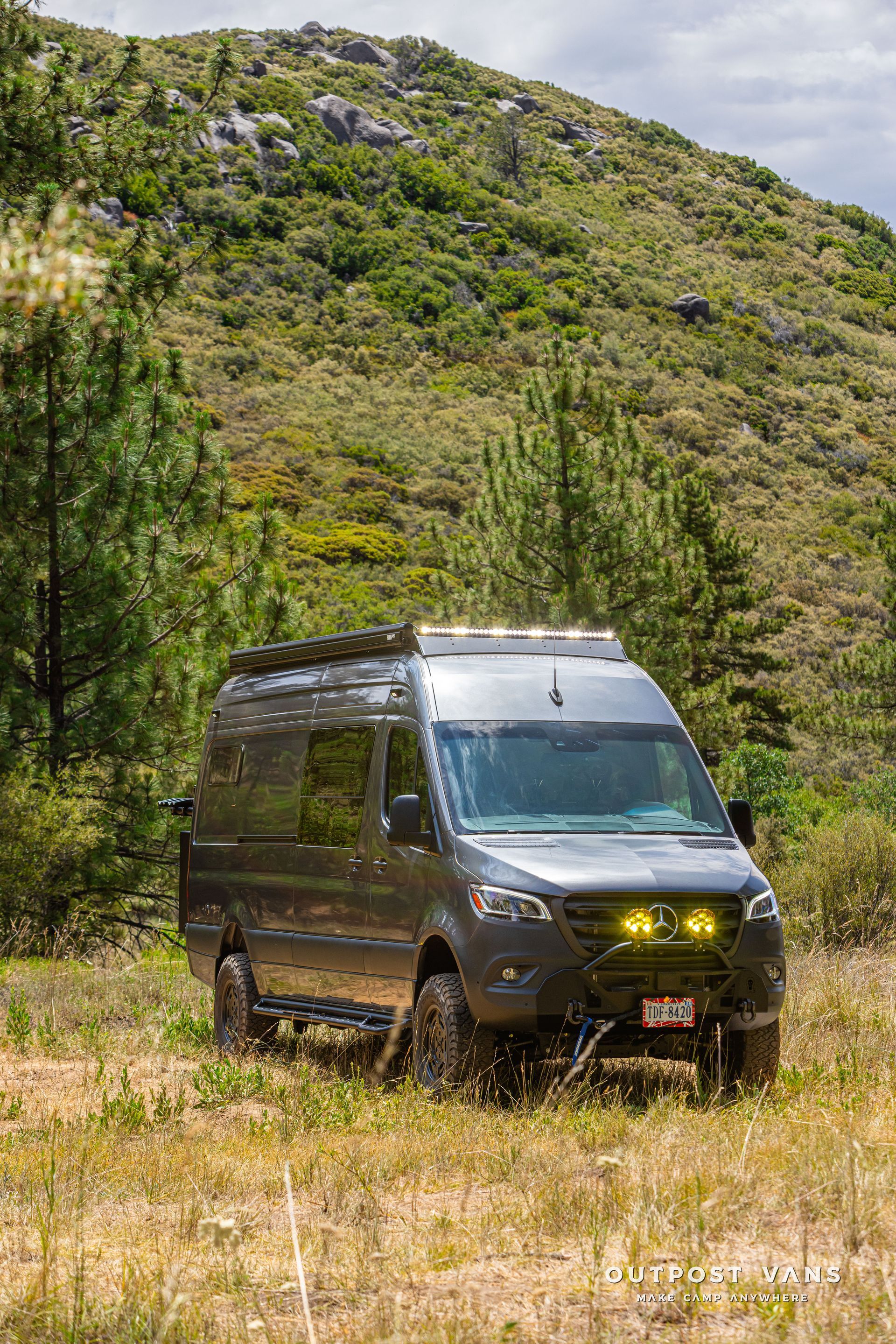 A van is parked in a grassy field in front of a mountain. Outpost vans Sprinter 170 AWD.