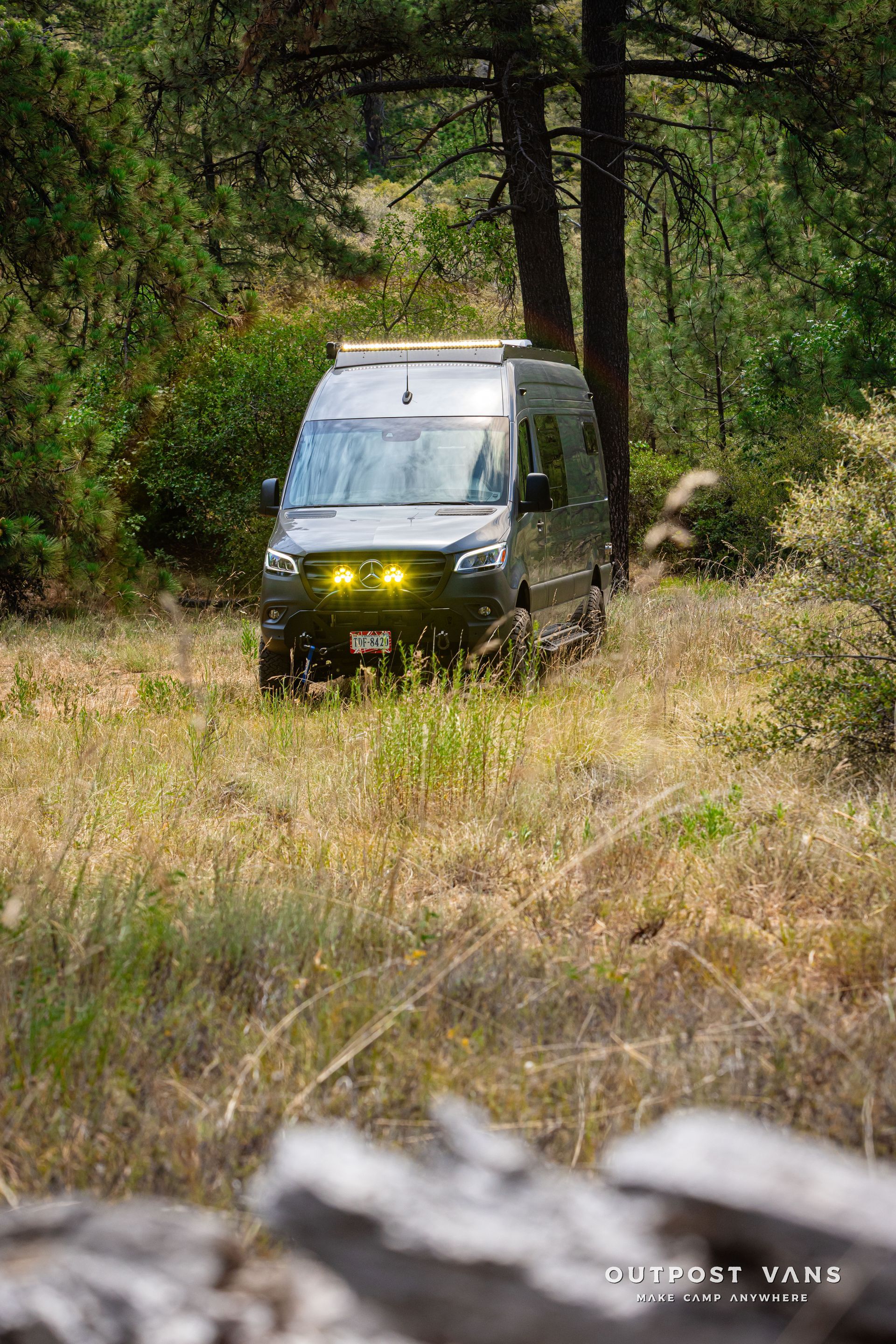 A van is driving through a grassy field in the woods. Outpost vans Sprinter 170 AWD.