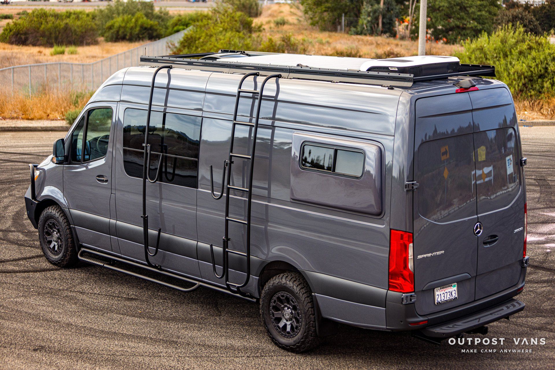 A gray van with a ladder on the roof is parked in a parking lot.