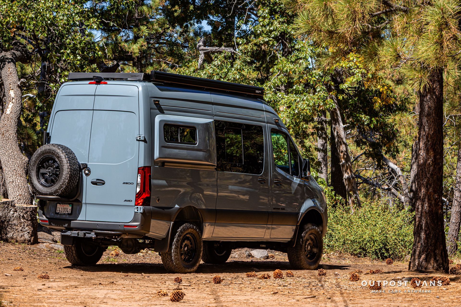 Sprinter camper van parked in a forest, spare tire on the back, dark wheels and roof rack.