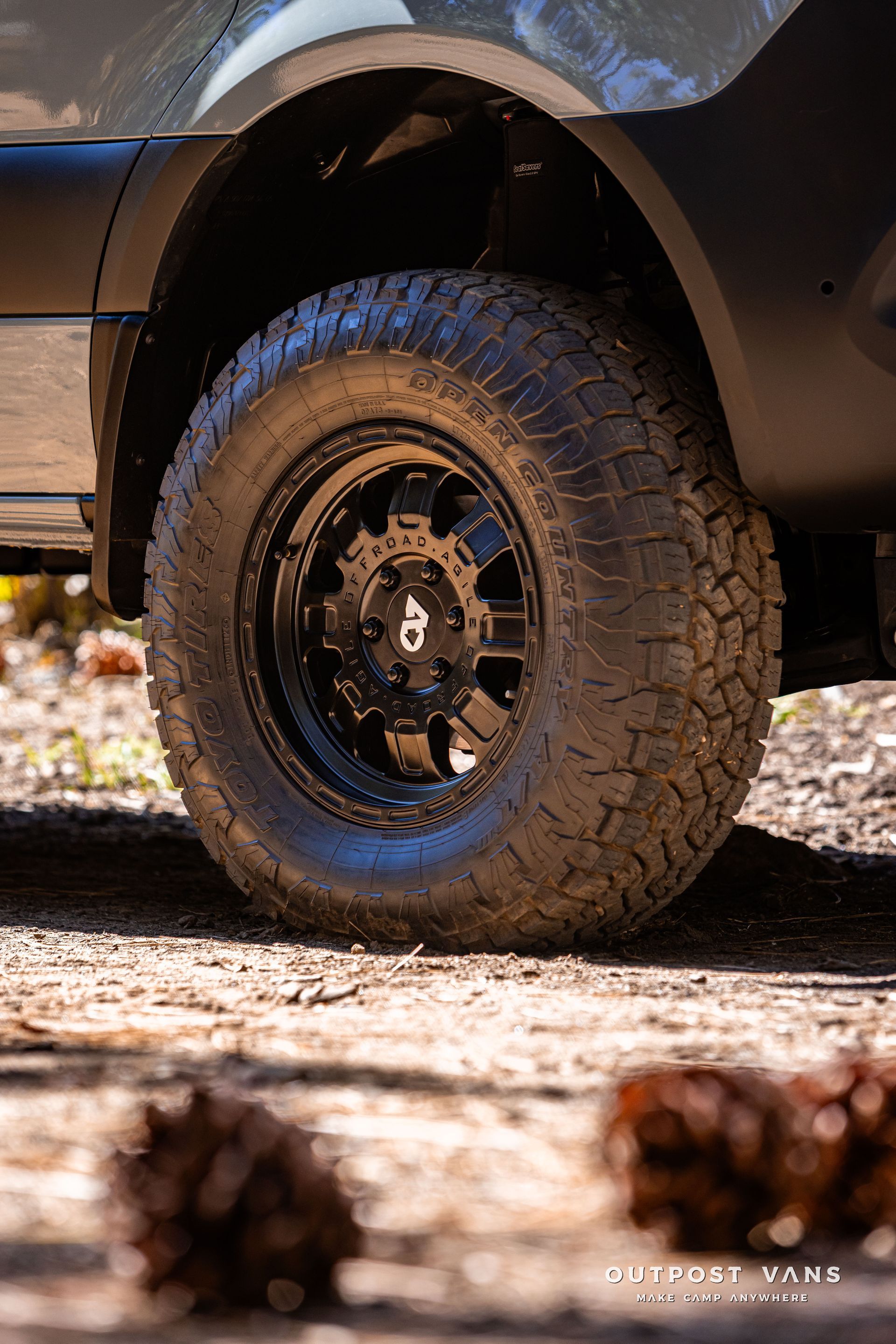 Sprinter Black off-road tire and wheel on a van, set in a forest scene with pine cones.