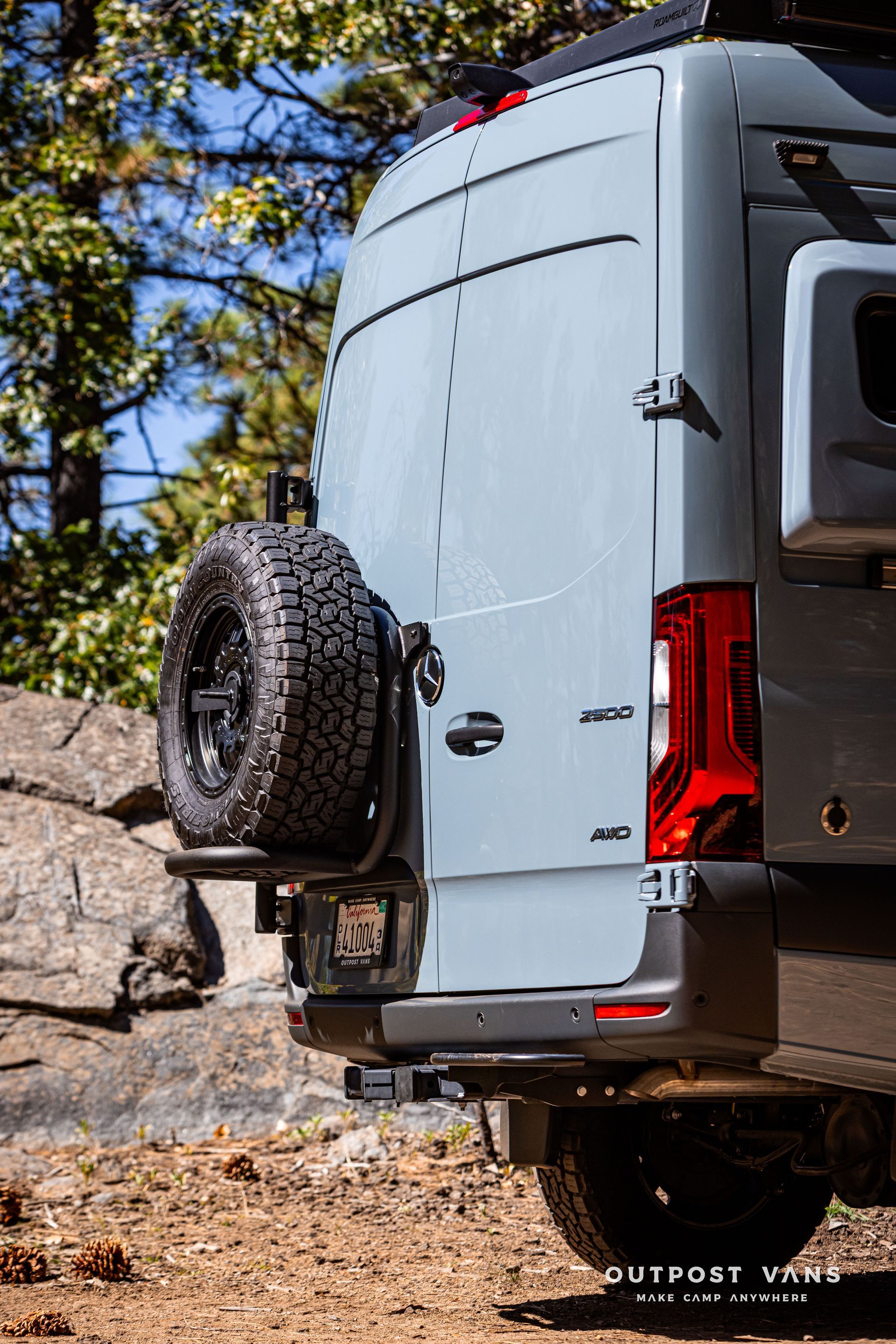 Sprinter Back of a gray adventure van with spare tire, in a forest setting.