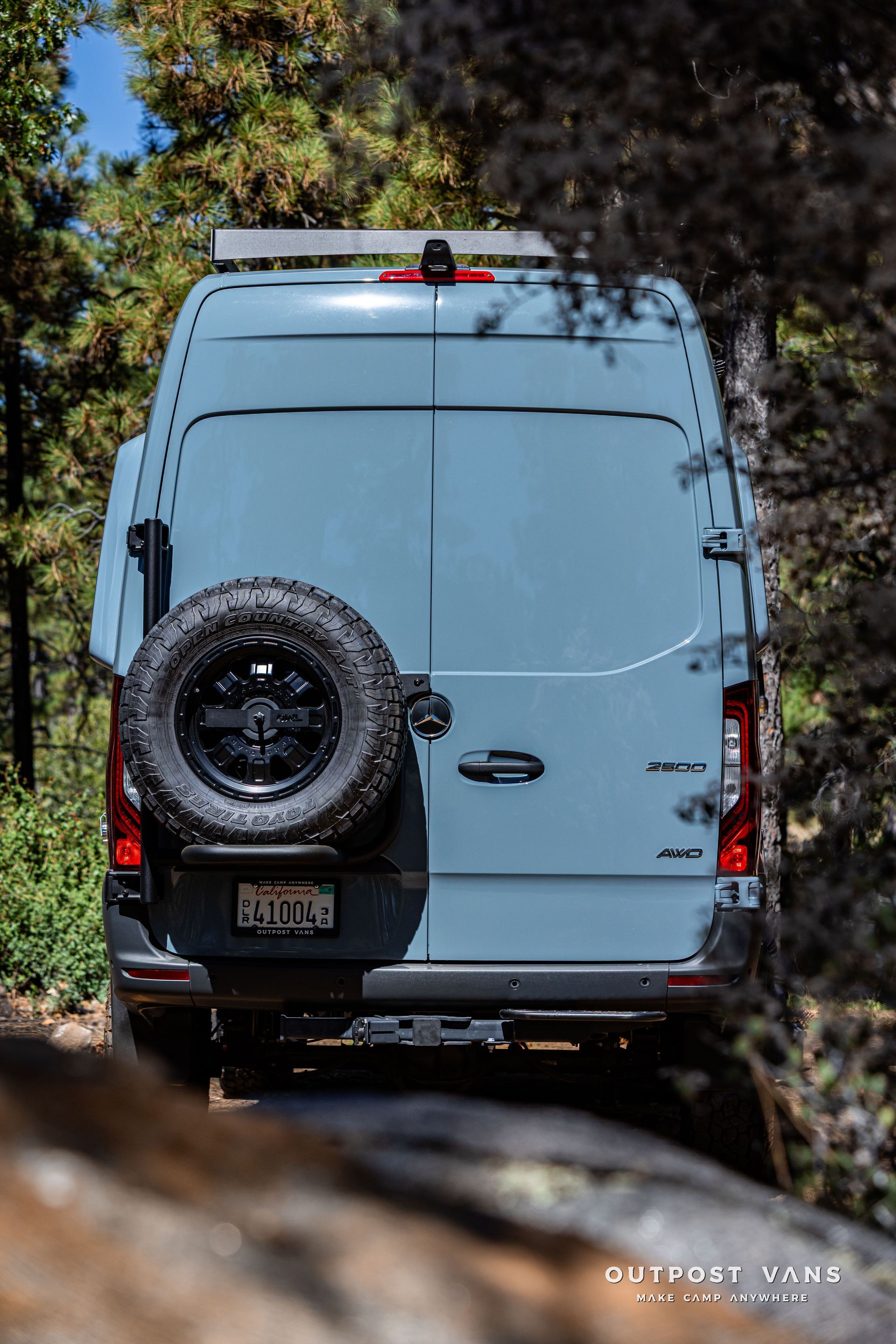 Sprinter Blue camper van parked on a dirt path in a forest, with a spare tire mounted on the back.
