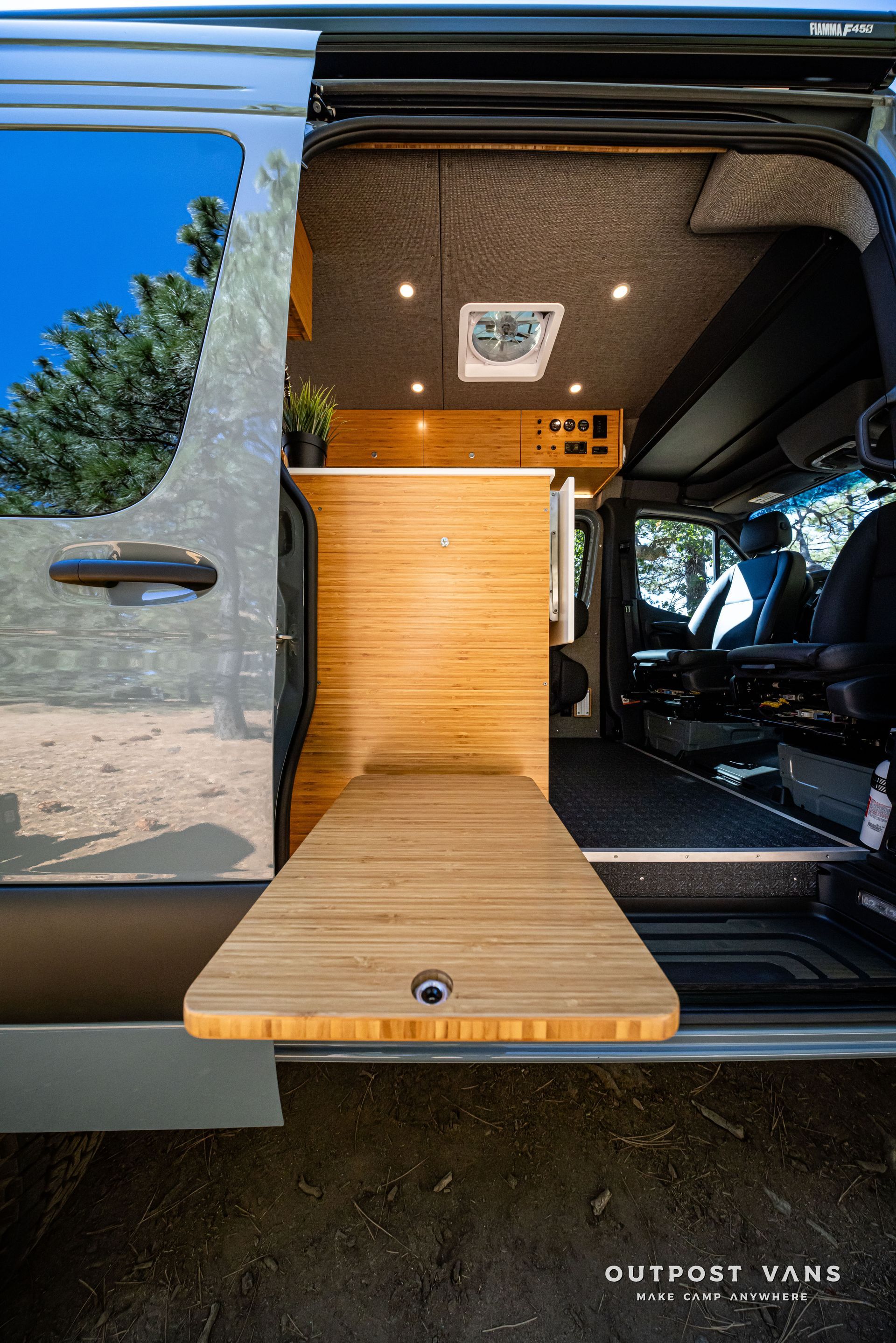SprinterVan interior with open door, extending wooden table. Bamboo cabinets, gray walls, and overhead lights.