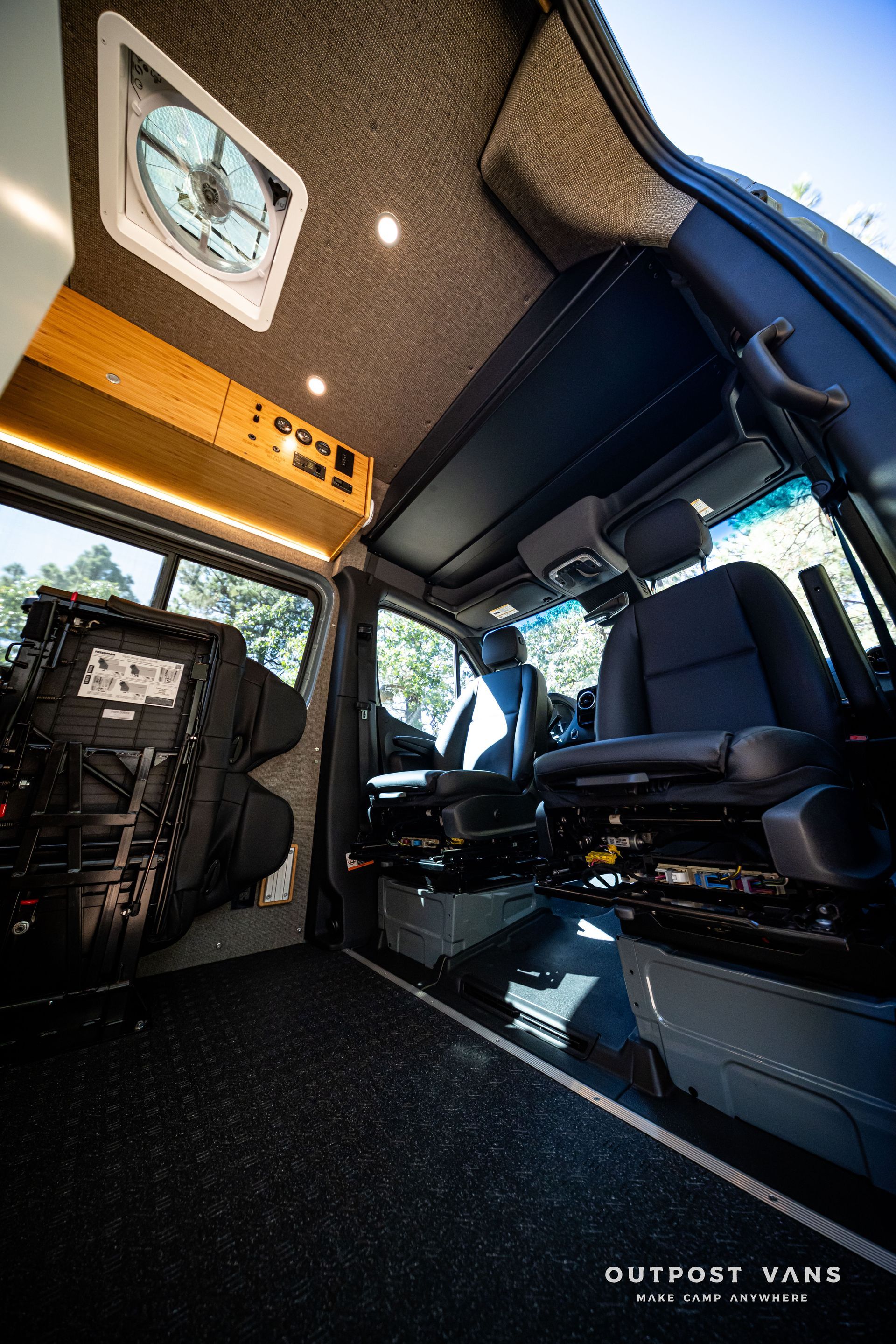 Sprinter Inside view of a converted van. Black seats, dark flooring, overhead vent, and storage against the wall.