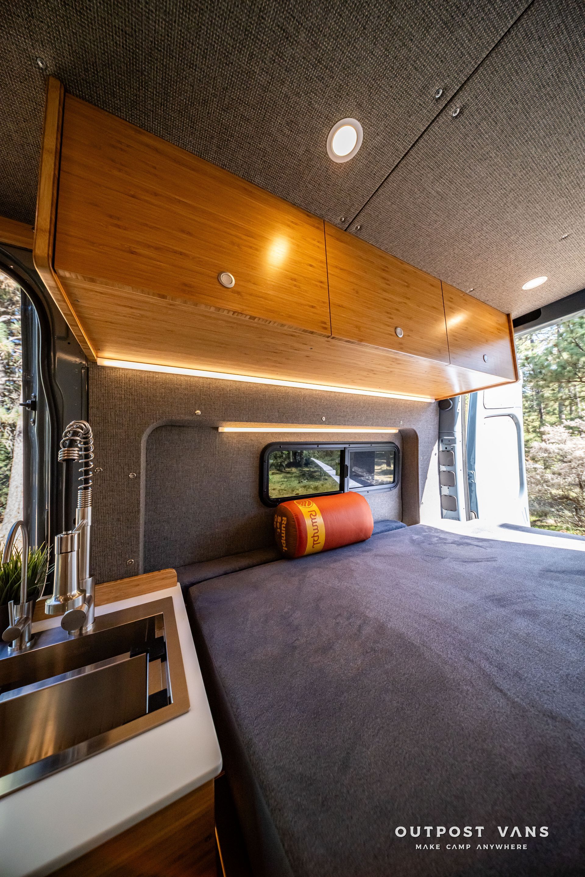 Sprinter Van interior with bed, overhead storage, and a sink. Gray and wood tones with accent lighting.