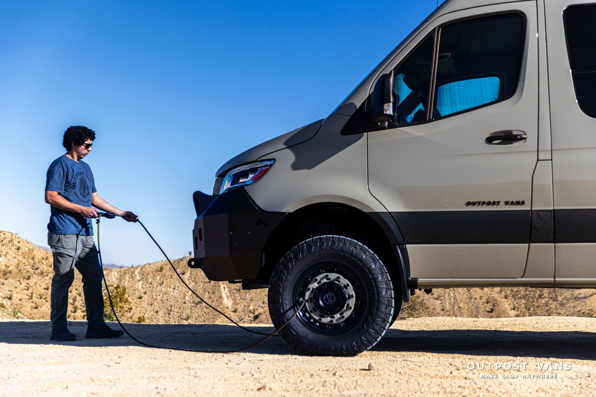 Person standing beside a beige van in a sandy desert, holding a tow rope attached to the front bumper.