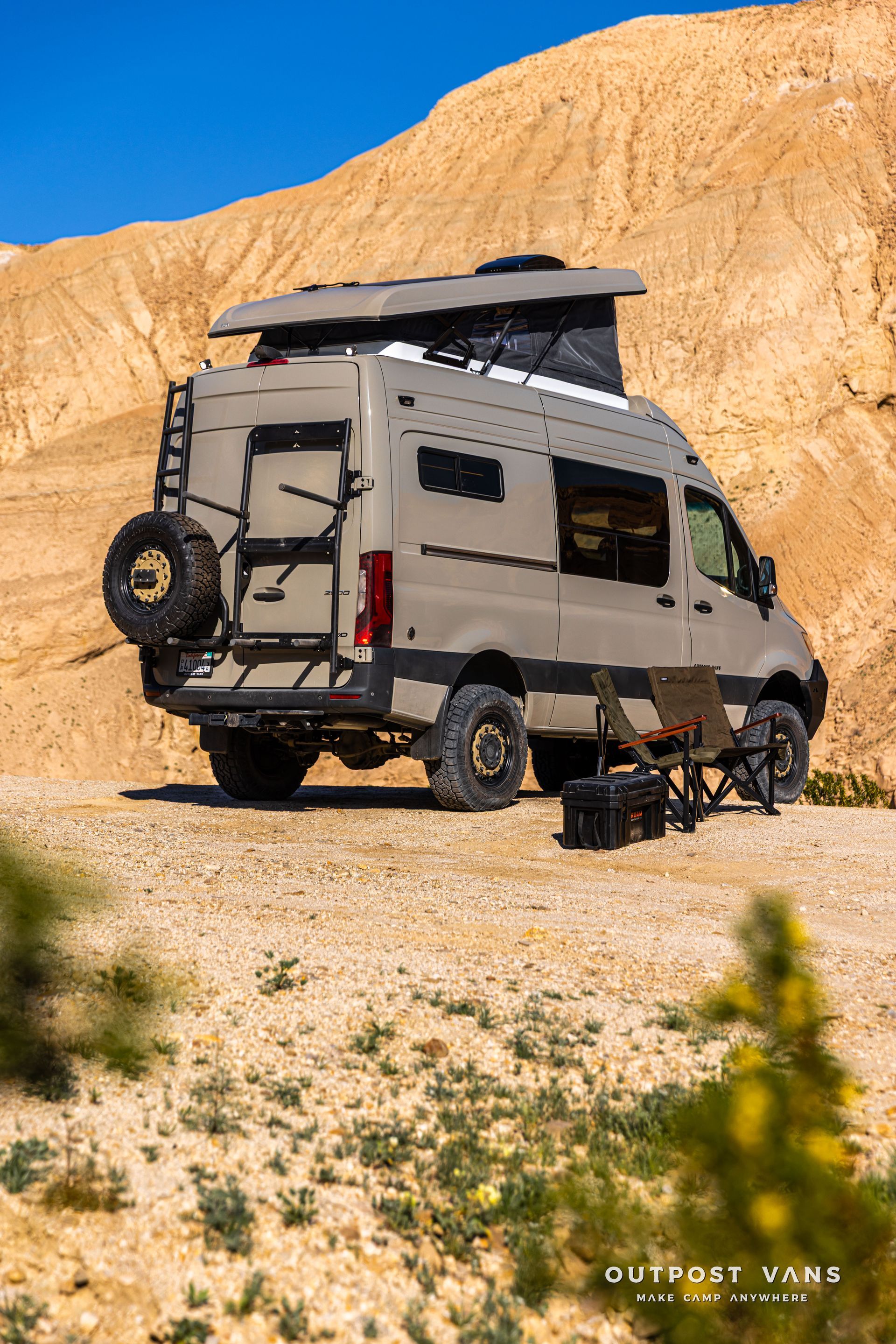 Off-road camper van parked on rocky desert terrain beside a tan cliff under a blue sky