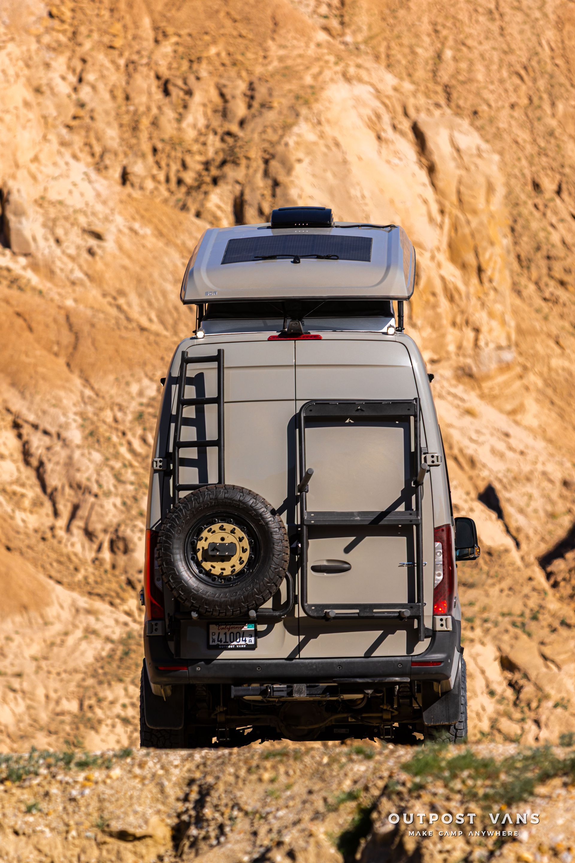 Gray van with rooftop cargo box and rear spare tire driving through a narrow rocky canyon.