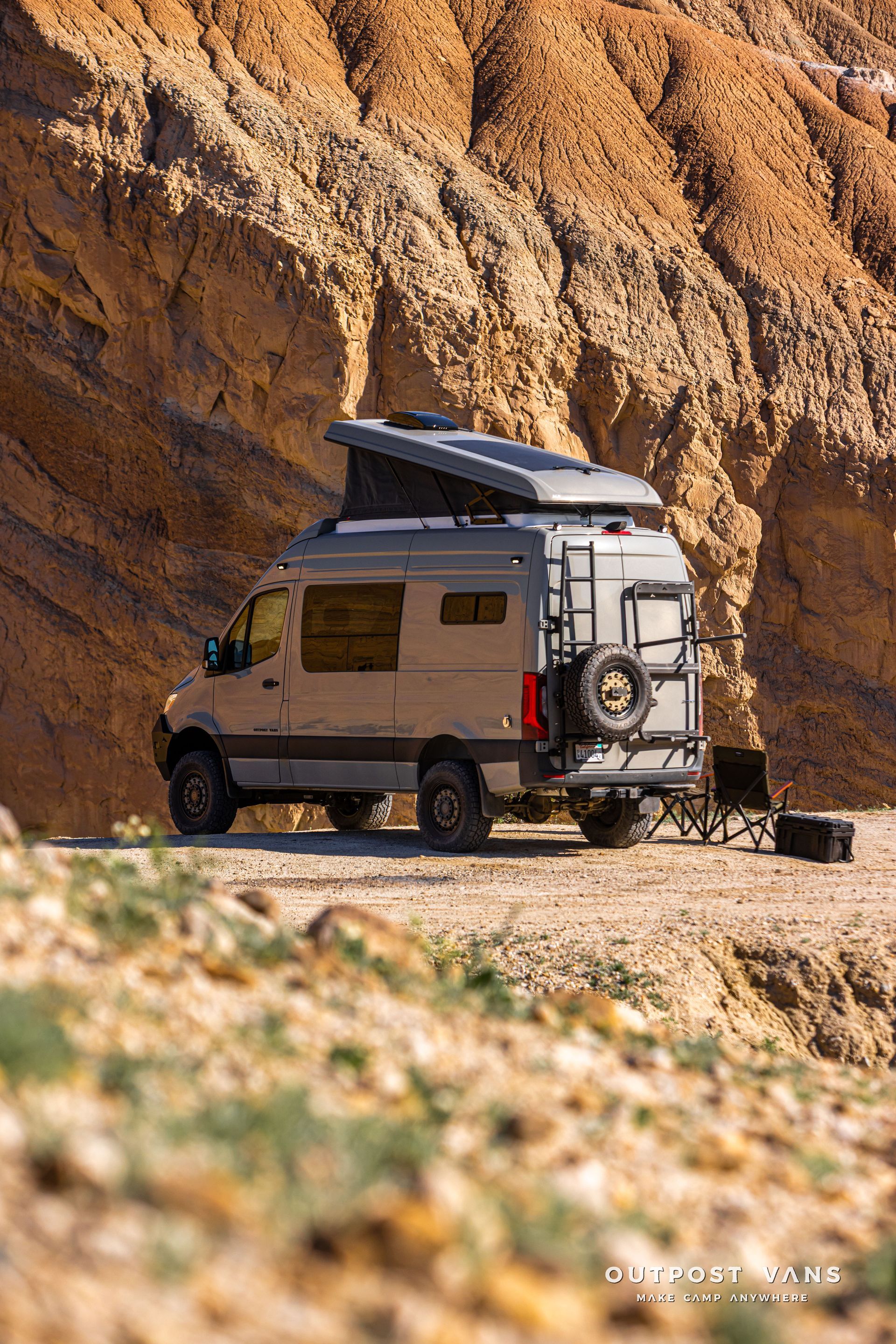 Off-road camper van parked beside a rocky canyon wall, with rooftop gear and a spare tire.